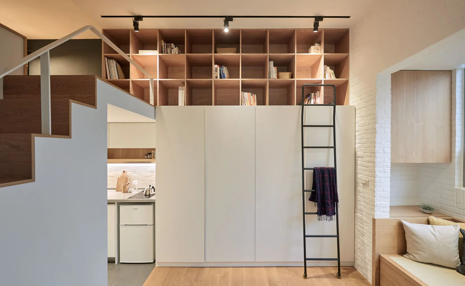 A modern micro-apartment interior features a white built-in storage cabinet topped with a wooden grid bookshelf and accessed by a black metal ladder.