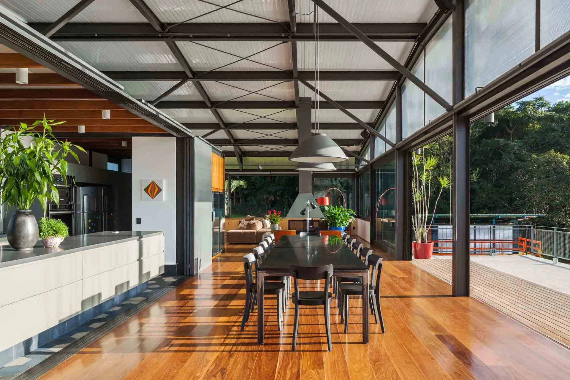 A modern open-plan dining area features a long black table, matching chairs, glossy wood floors, and exposed dark steel roof trusses next to a deck.