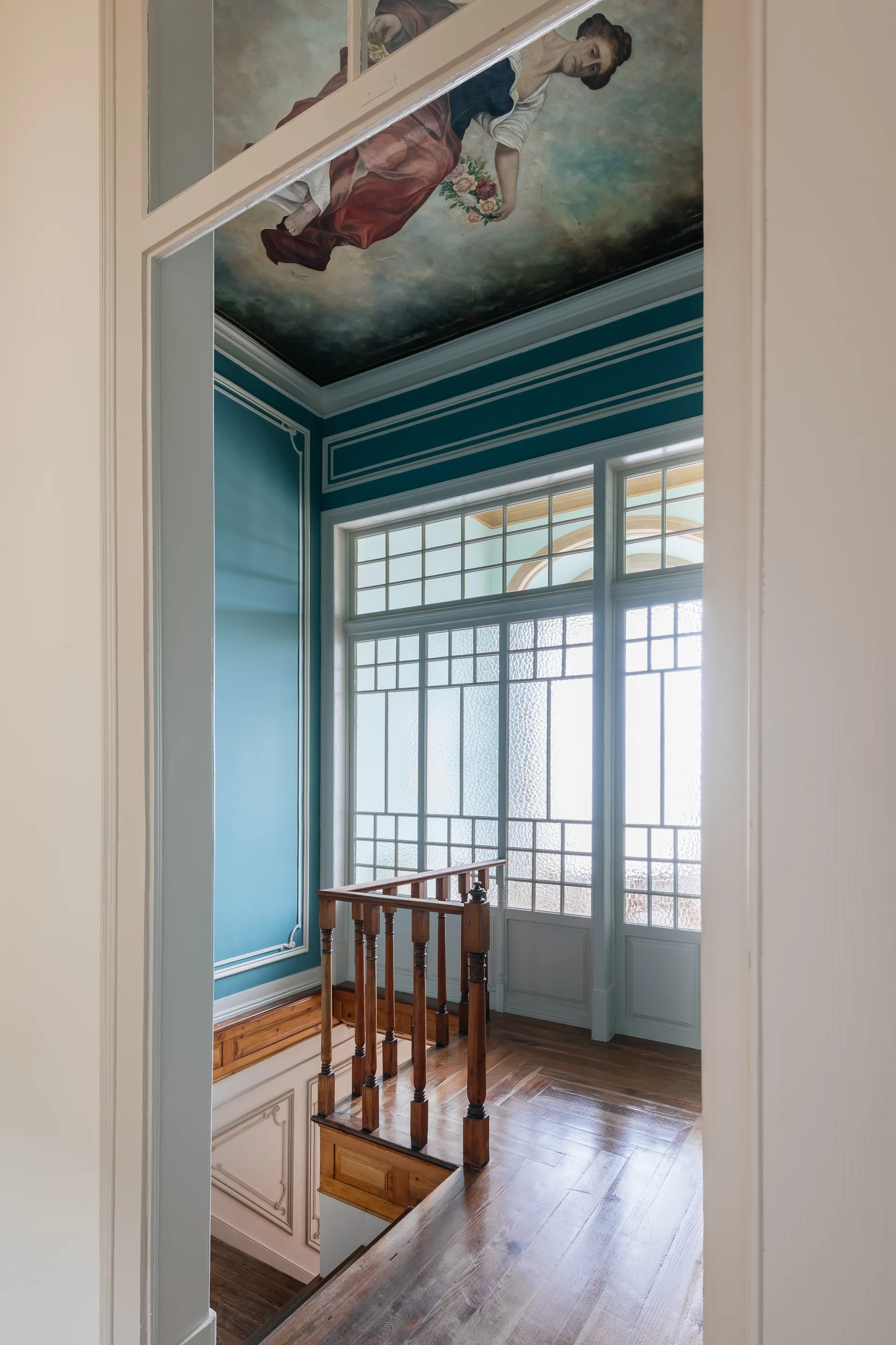 An interior staircase landing features teal walls with white molding, a natural wood balustrade, a frosted glass partition, and a painted ceiling mural.