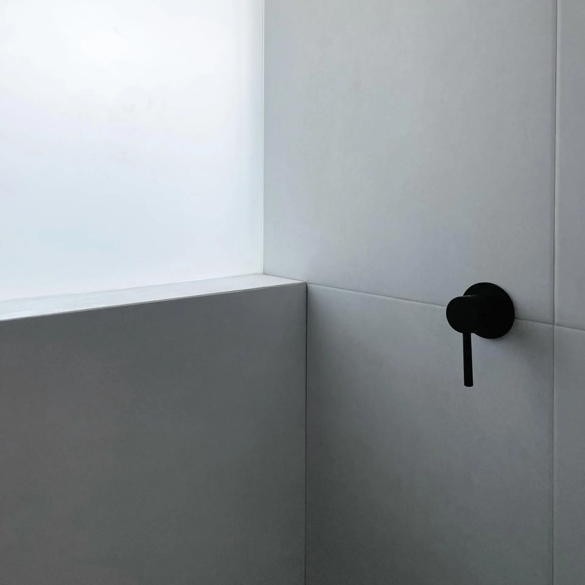 A close-up of a minimalist matte black faucet handle mounted on light grey large-format wall tiles next to a frosted glass window.