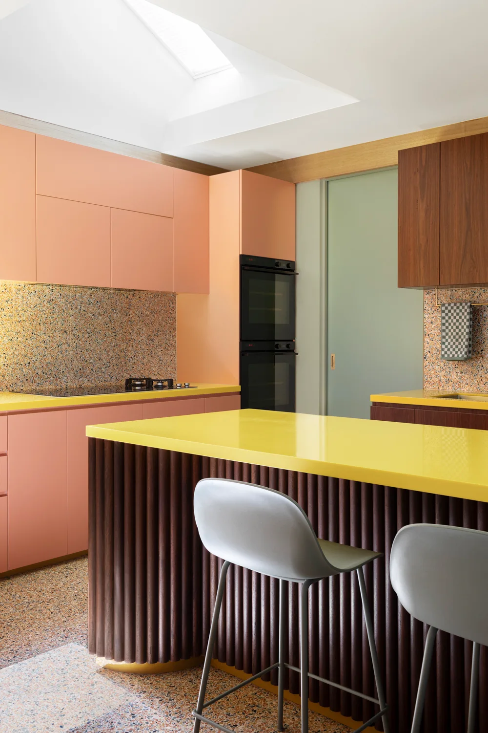 A modern kitchen featuring a dark fluted wood island with a yellow countertop, pink cabinets, and speckled terrazzo flooring.