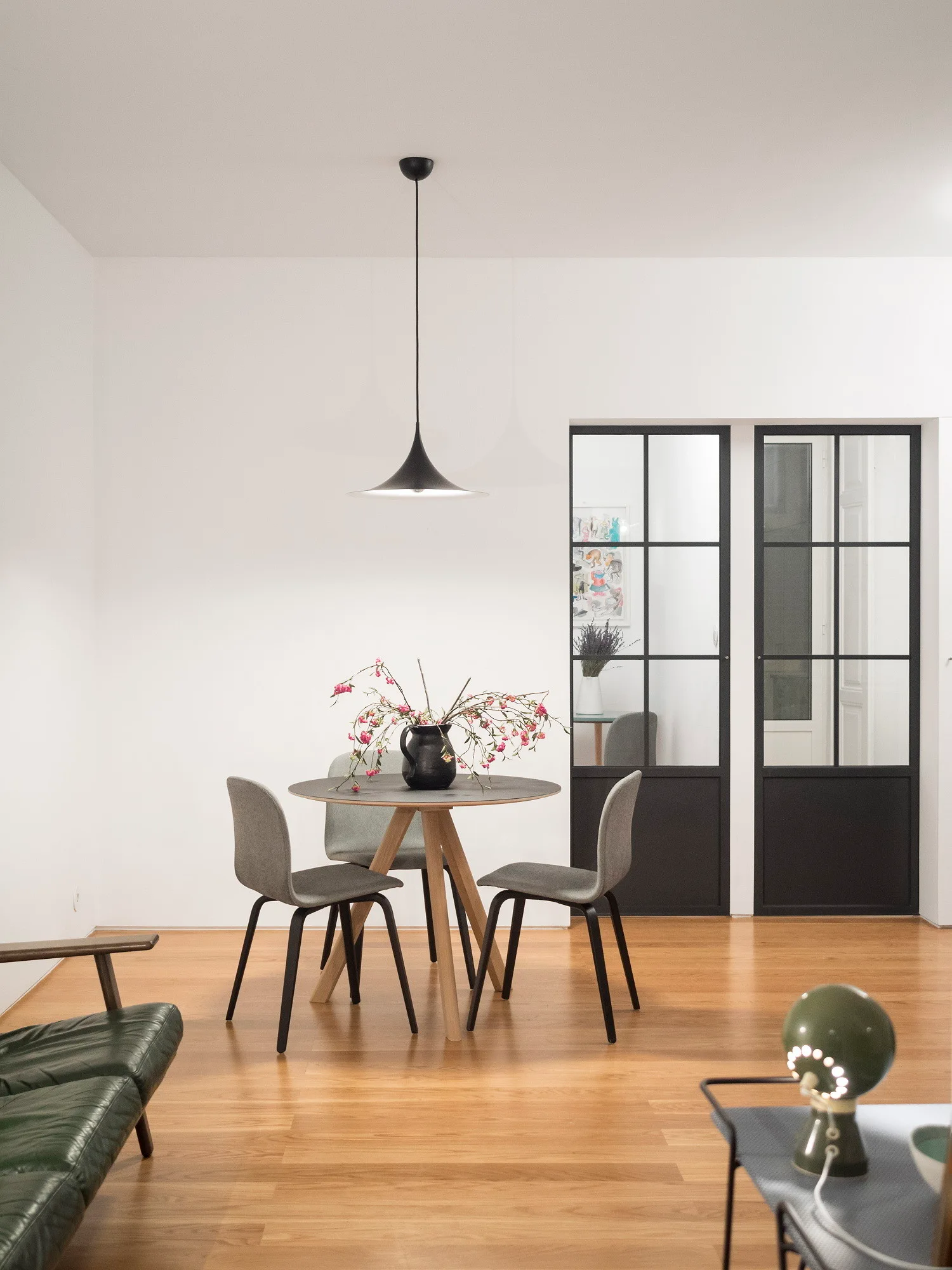 A minimalist dining area features a round wooden table, grey chairs, a black cone pendant light, and black-framed glass French doors.