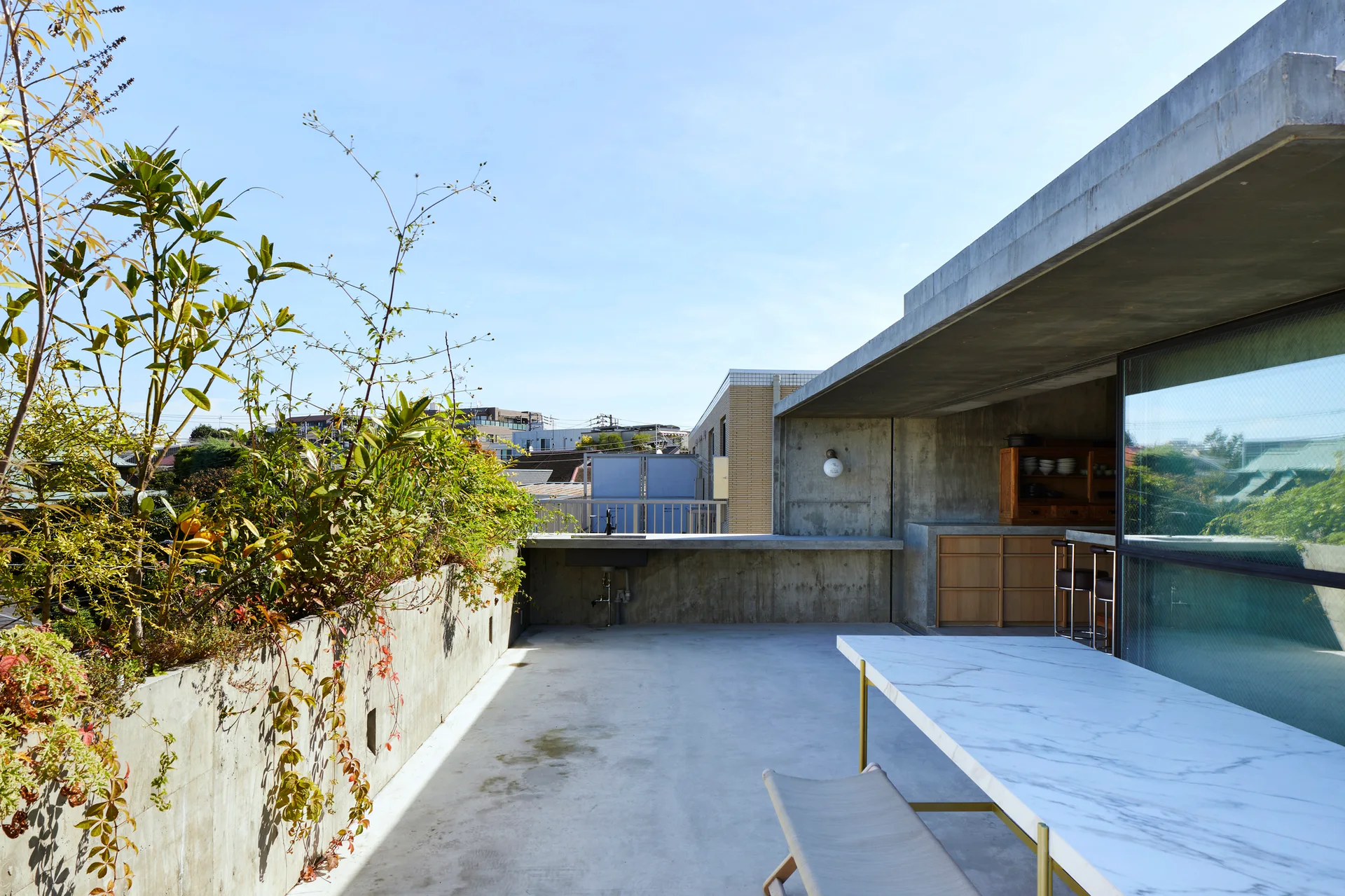 A concrete rooftop terrace features a large marble-topped dining table, an integrated concrete planter with green foliage, and built-in wood cabinetry.