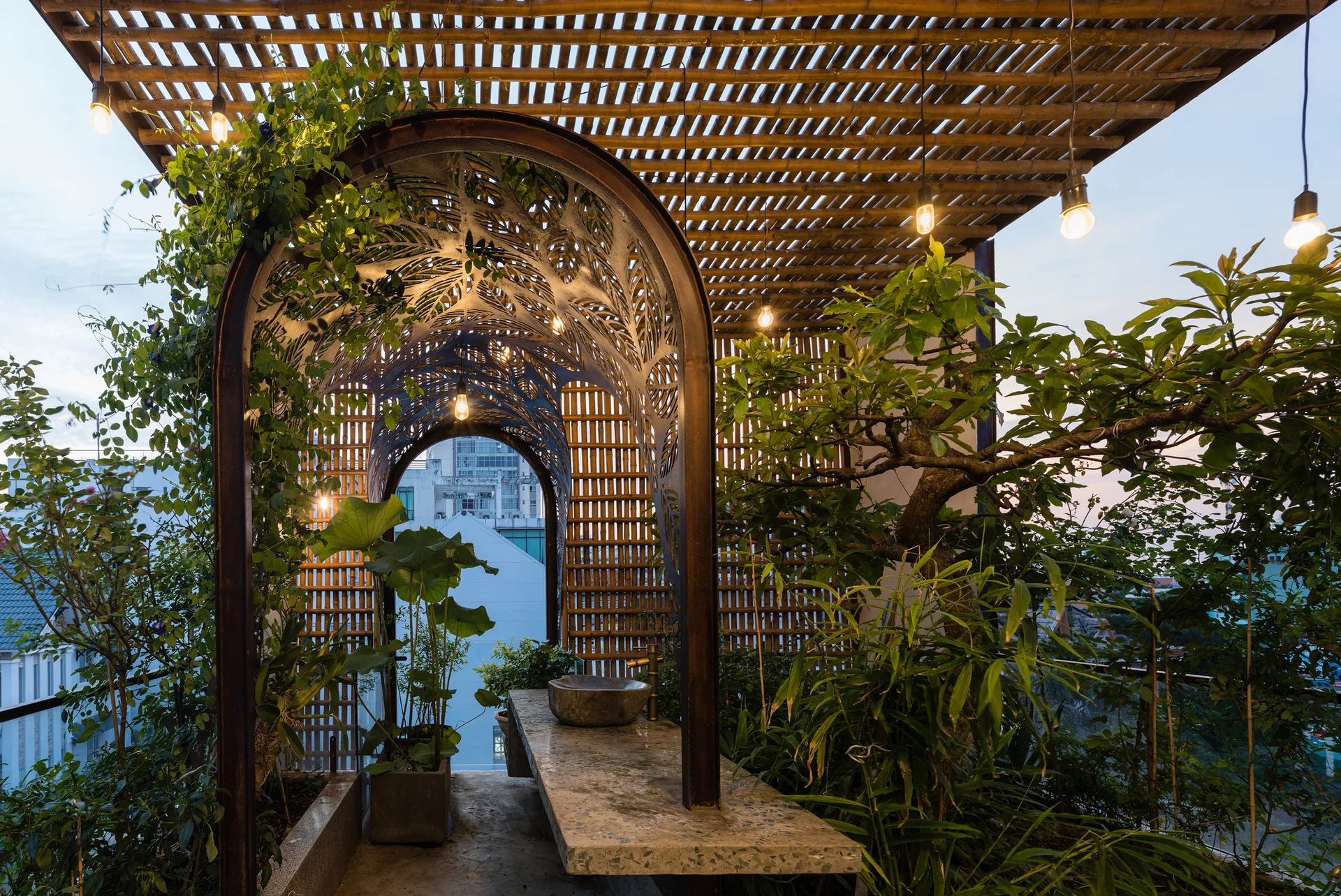 A rooftop terrace features an ornate metal archway over a stone vanity with a vessel sink, surrounded by lush plants under a slatted bamboo ceiling.