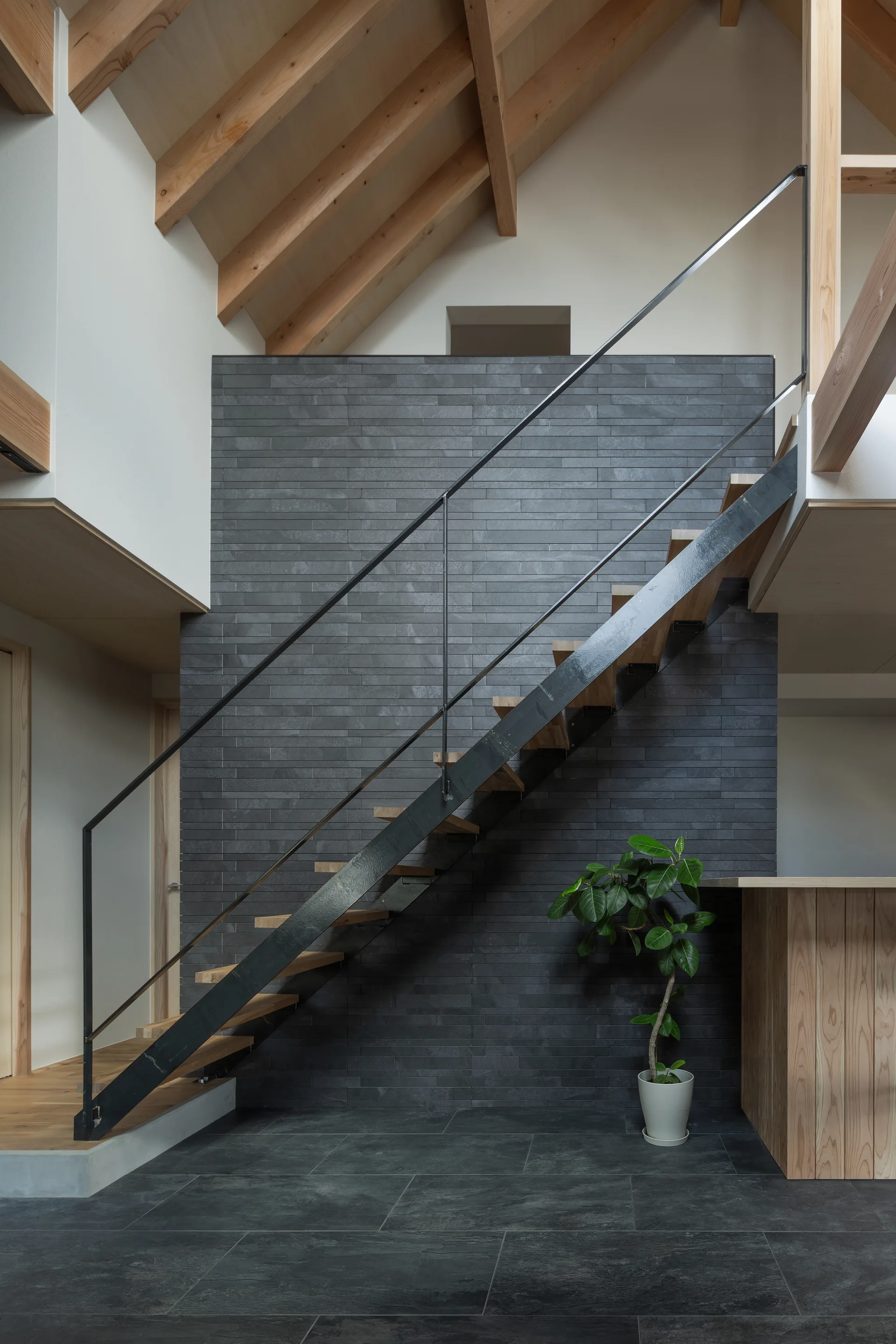 A minimalist floating wood staircase with a black metal railing stands against a dark gray tiled accent wall under exposed timber ceiling beams.