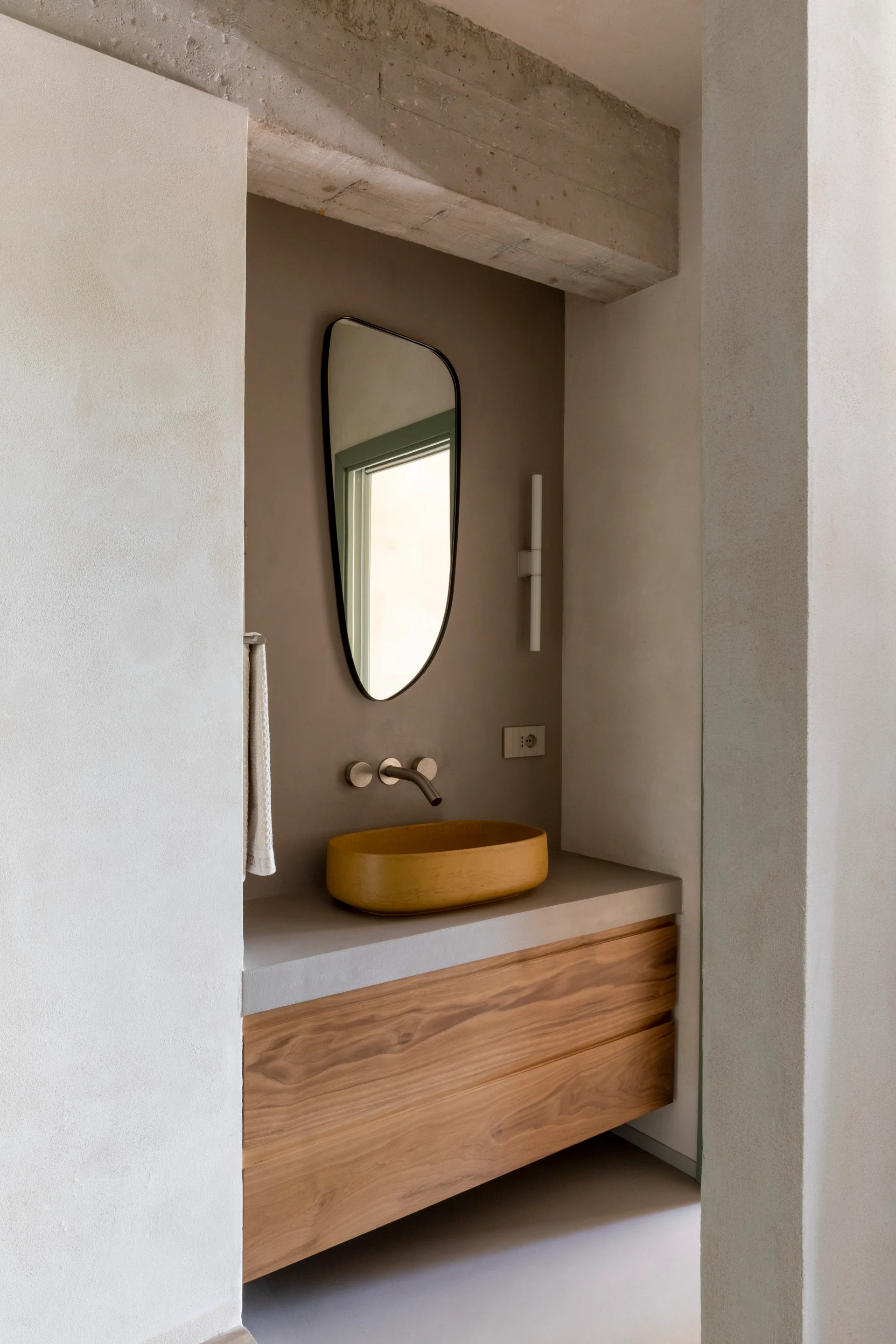 A minimalist bathroom alcove features a wooden floating vanity, a yellow vessel sink, an asymmetrical mirror, and an exposed concrete beam.