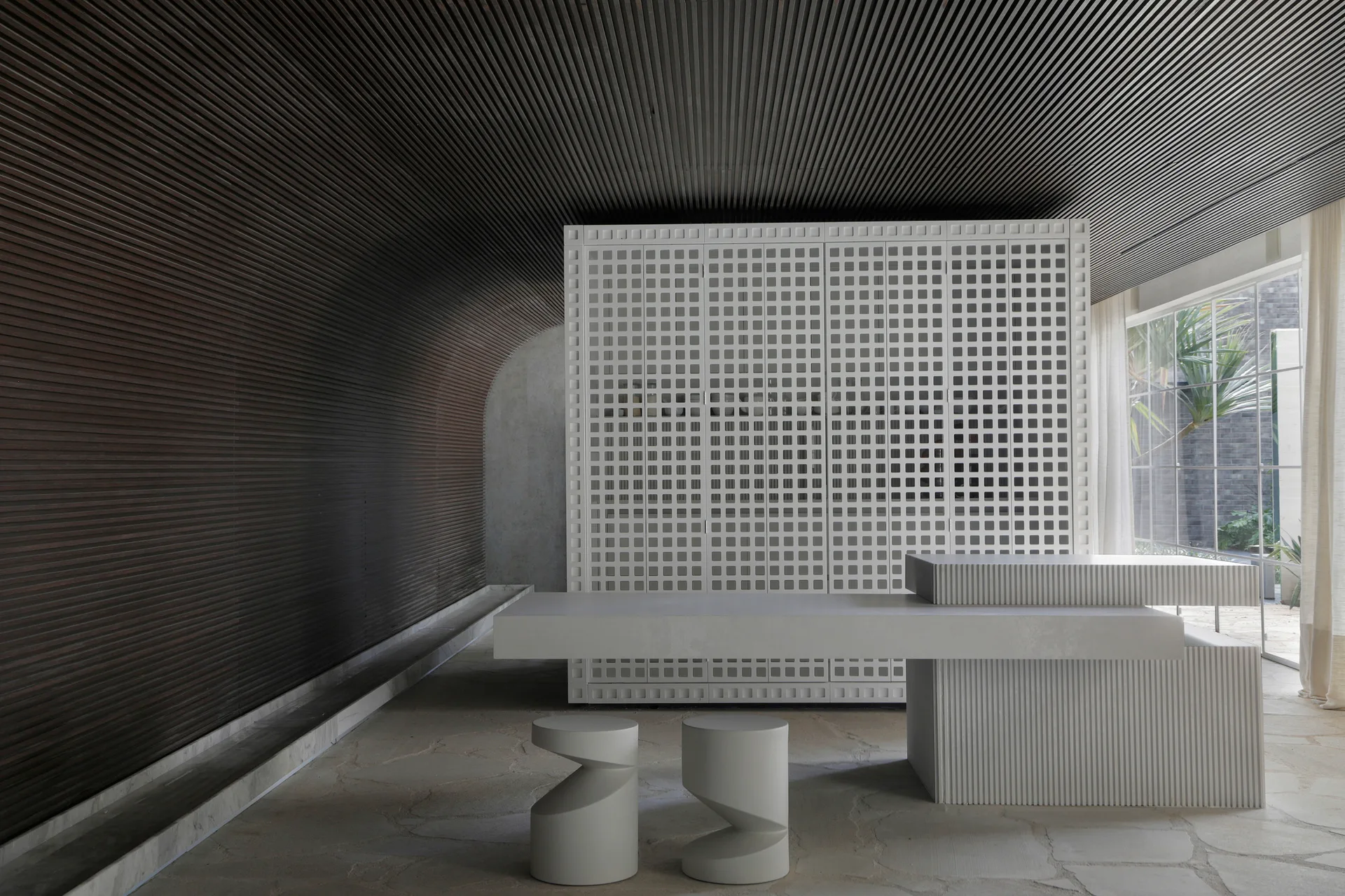 A minimalist interior featuring a dark slatted wood curved wall, a white grid cabinet, a long white table, and grey flagstone flooring.
