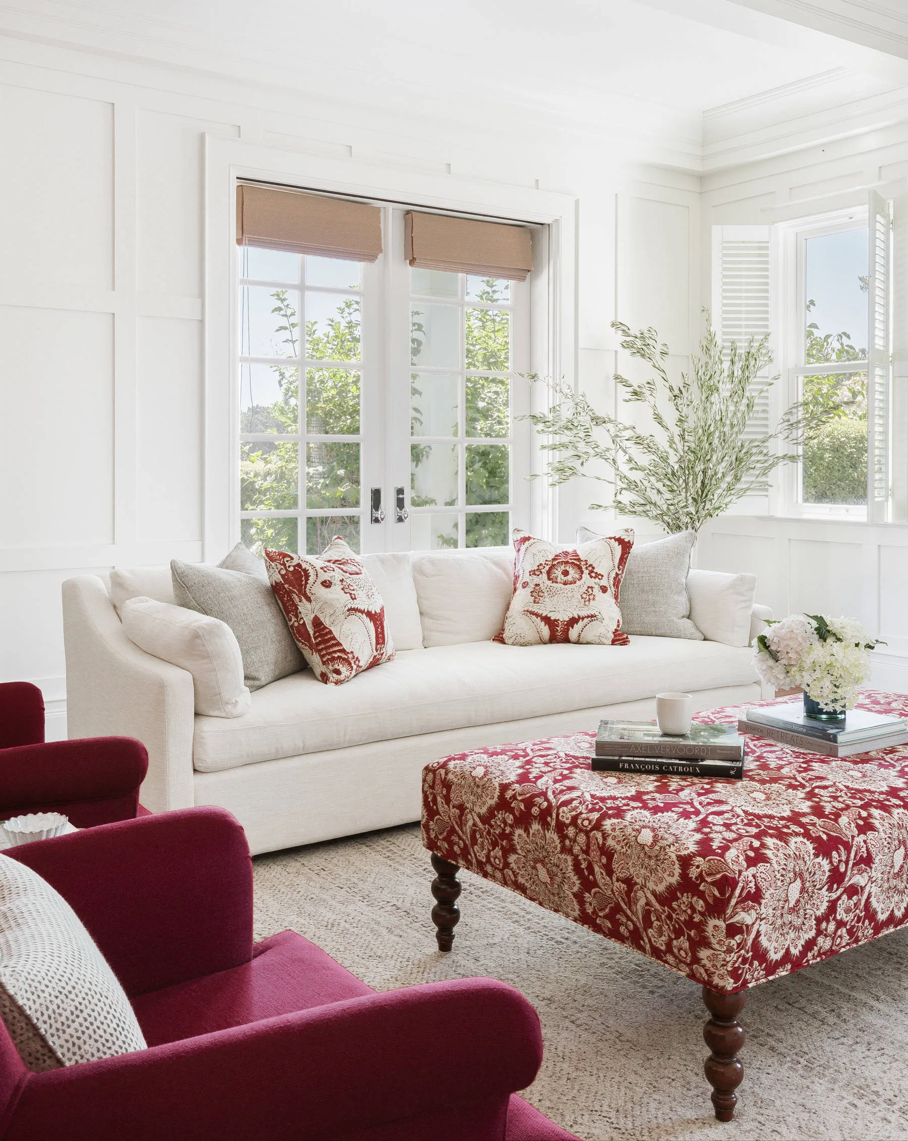 A bright living room features a white sofa, deep red armchairs, and a large red patterned ottoman set against white grid paneled walls.