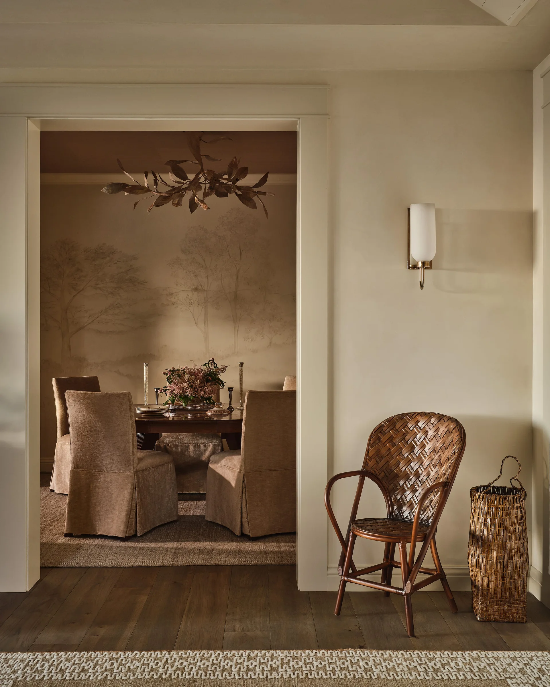 A view through a doorway into a dining room featuring a scenic tree mural, upholstered slipcovered chairs, and a branch chandelier.
