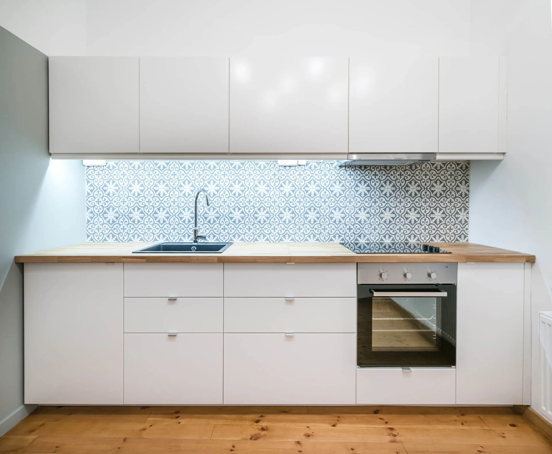 A modern kitchen with flat-front white cabinets, a light wood countertop, a blue and white patterned tile backsplash, and built-in appliances.