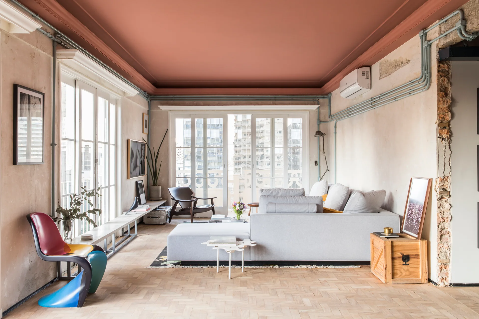 A spacious living room featuring a terracotta painted ceiling, distressed plaster walls with exposed blue conduit, and a gray sectional sofa.