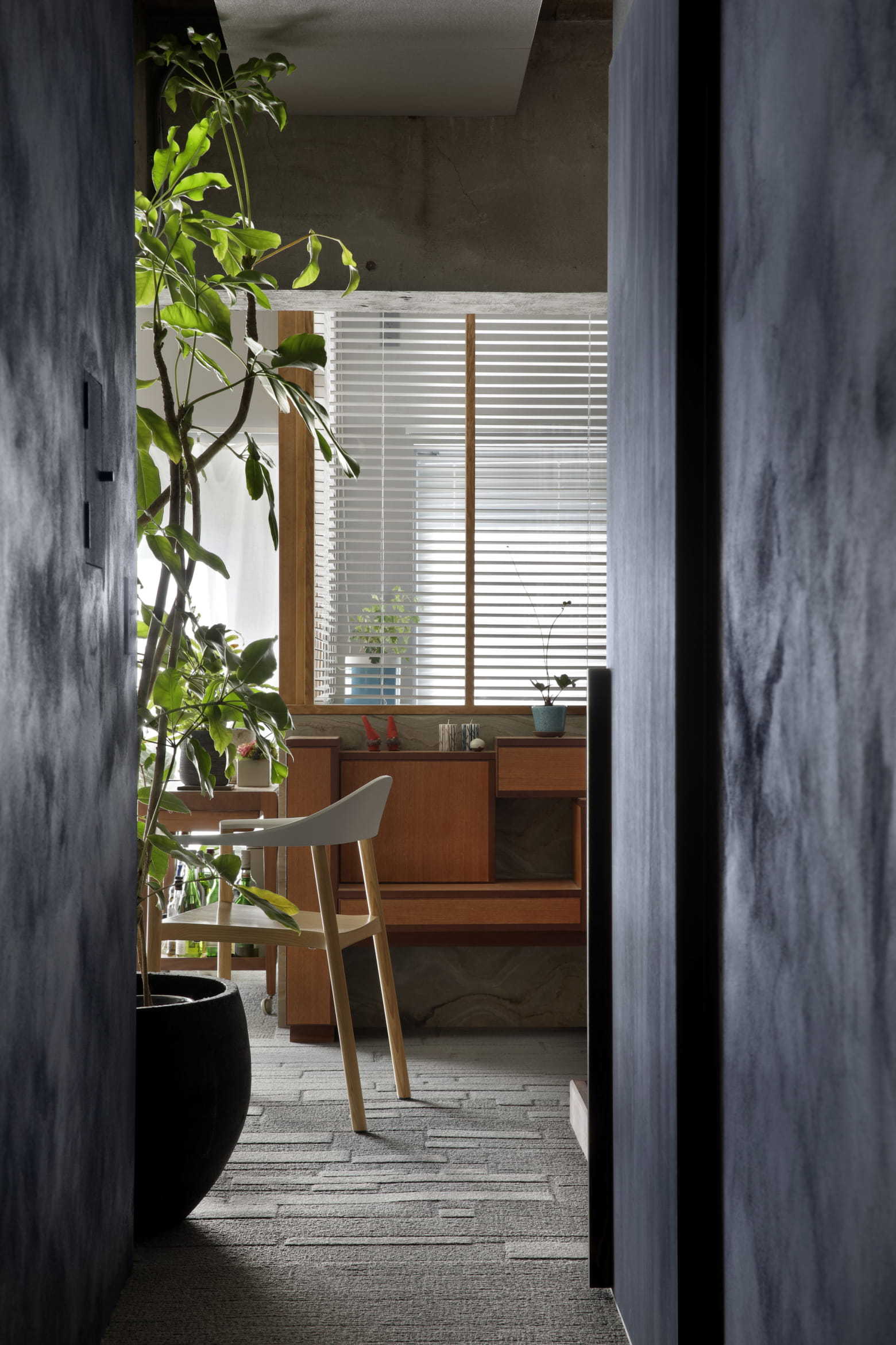 A view through a dark-toned doorway reveals a wooden console cabinet, a minimalist chair, window blinds, and a large potted plant in the foreground.