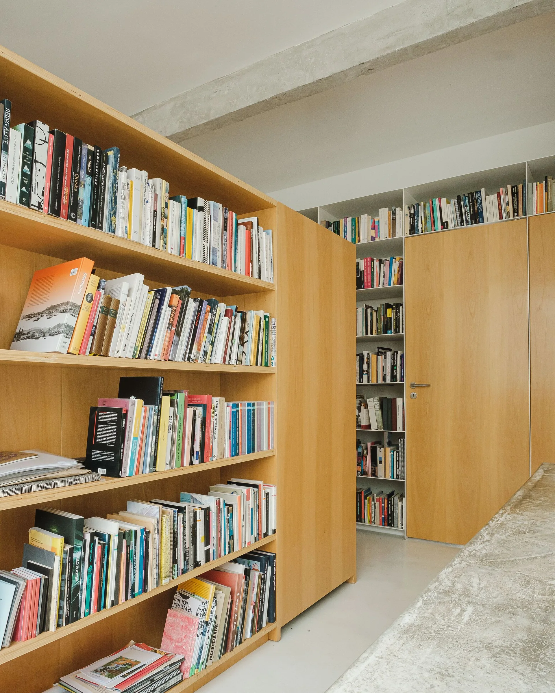 Light wood floor-to-ceiling bookshelves filled with books surround a matching wooden door beneath an exposed concrete ceiling beam.
