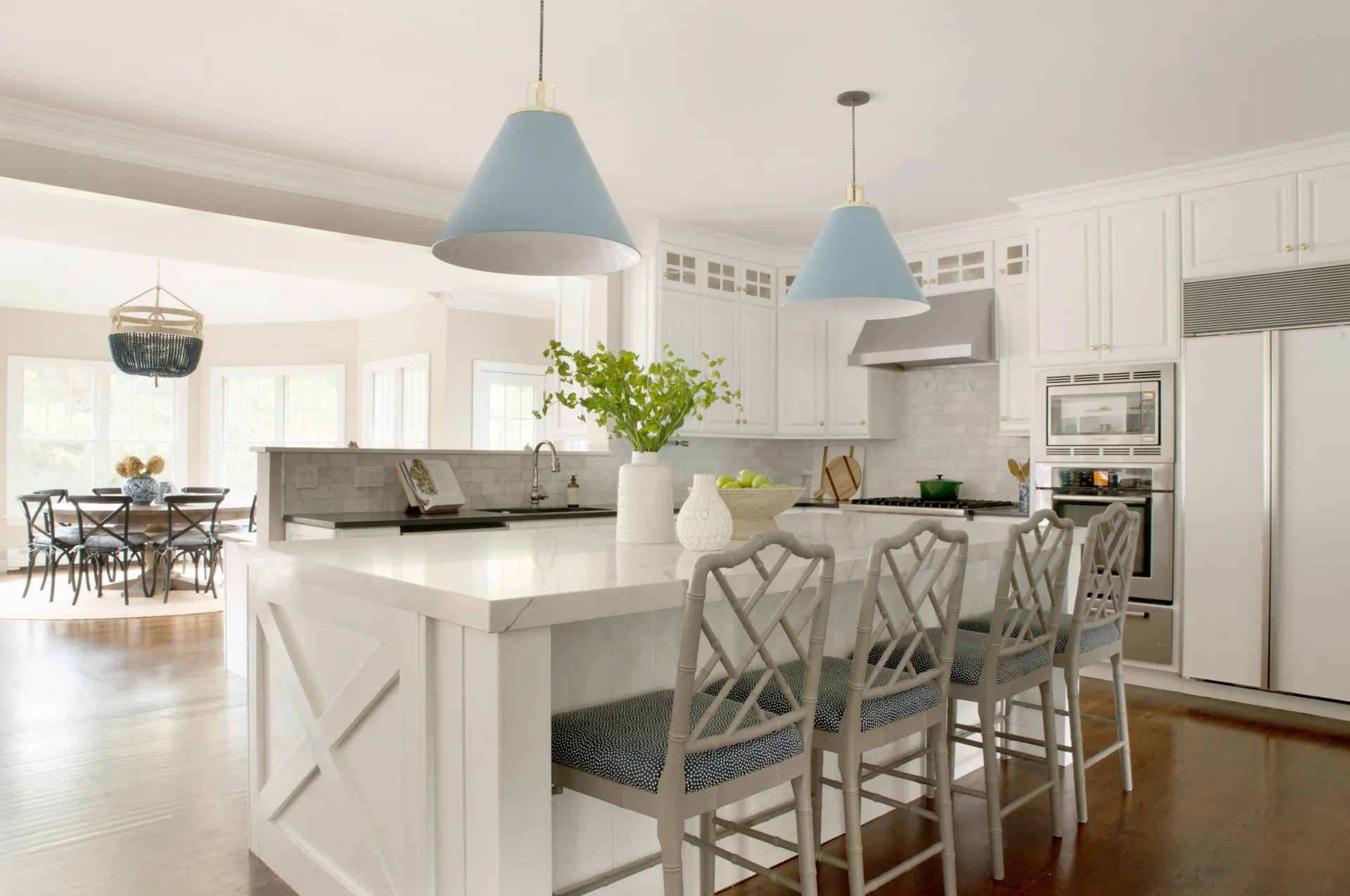 A bright white kitchen featuring a large central island with lattice-back bar stools, two blue dome pendant lights, and stainless steel appliances.