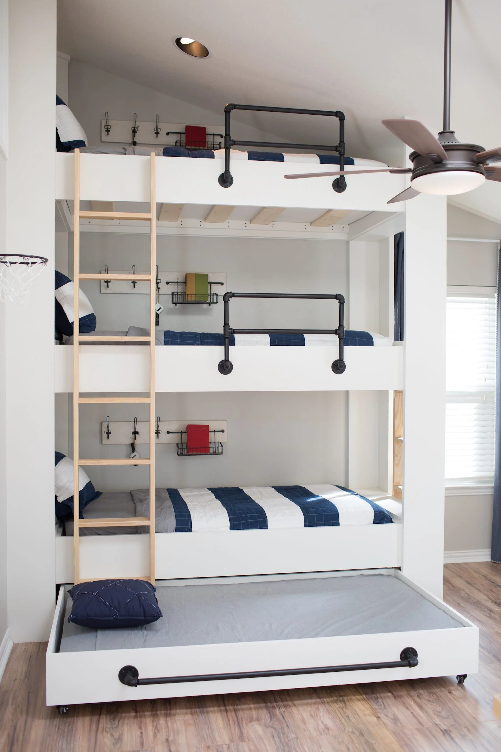 A built-in white triple bunk bed features a light wood ladder, black pipe safety rails, striped bedding, and a pulled-out bottom trundle bed.