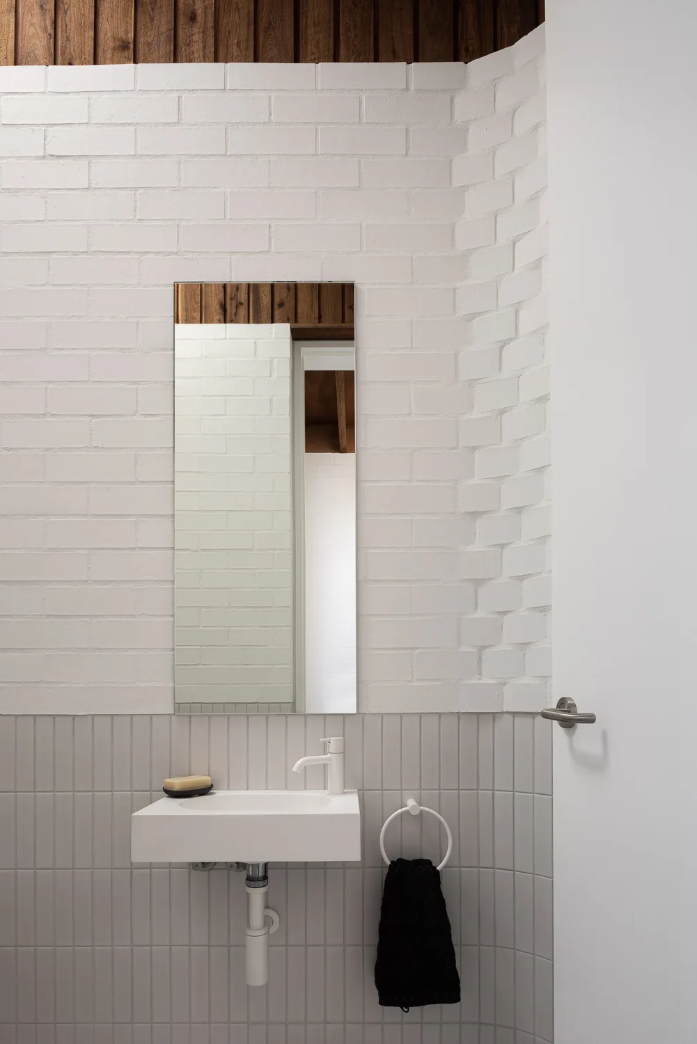 A minimalist powder room features a white wall-mounted sink below a frameless mirror against painted brick and grey vertically stacked tiles.
