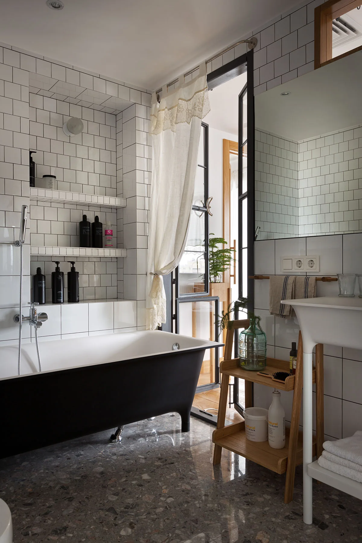A modern bathroom features a black freestanding bathtub, white grid-tiled walls, a pedestal sink, and polished grey terrazzo flooring.