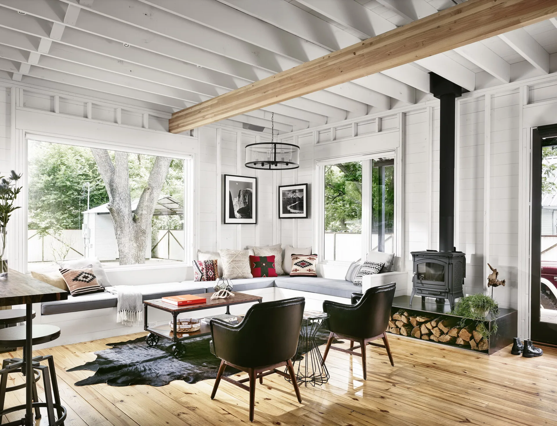 A bright white cabin living room featuring a corner built-in window bench, a black wood-burning stove with log storage, and exposed ceiling beams.