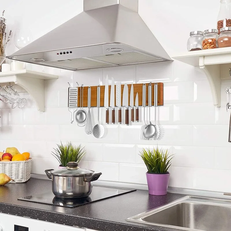 A wooden magnetic knife rack holding knives and kitchen utensils is mounted on a white subway tile backsplash above a dark countertop.