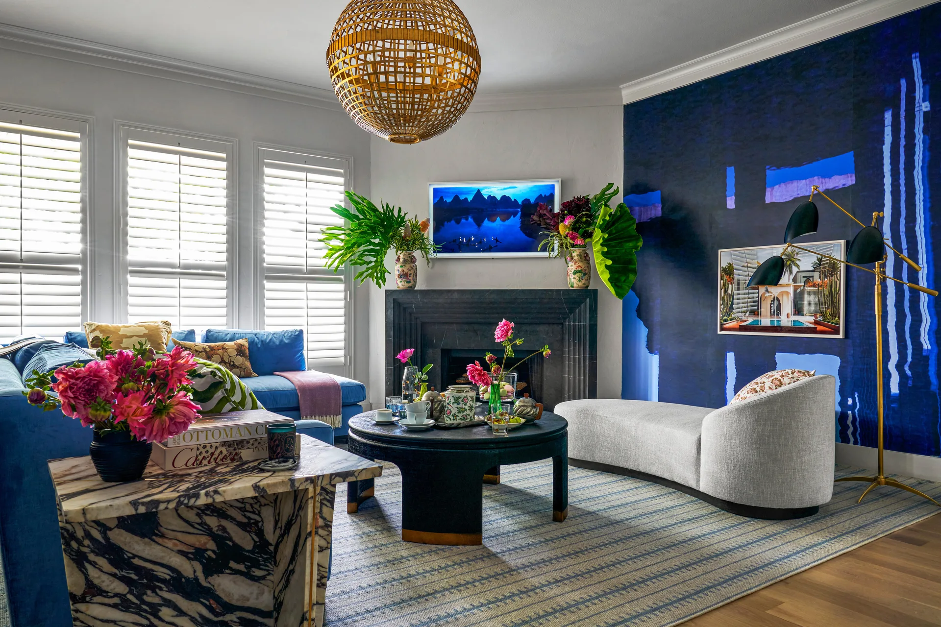 A living room featuring a blue sofa, light grey chaise, black stone fireplace, and a glossy deep blue accent wall next to shuttered windows.