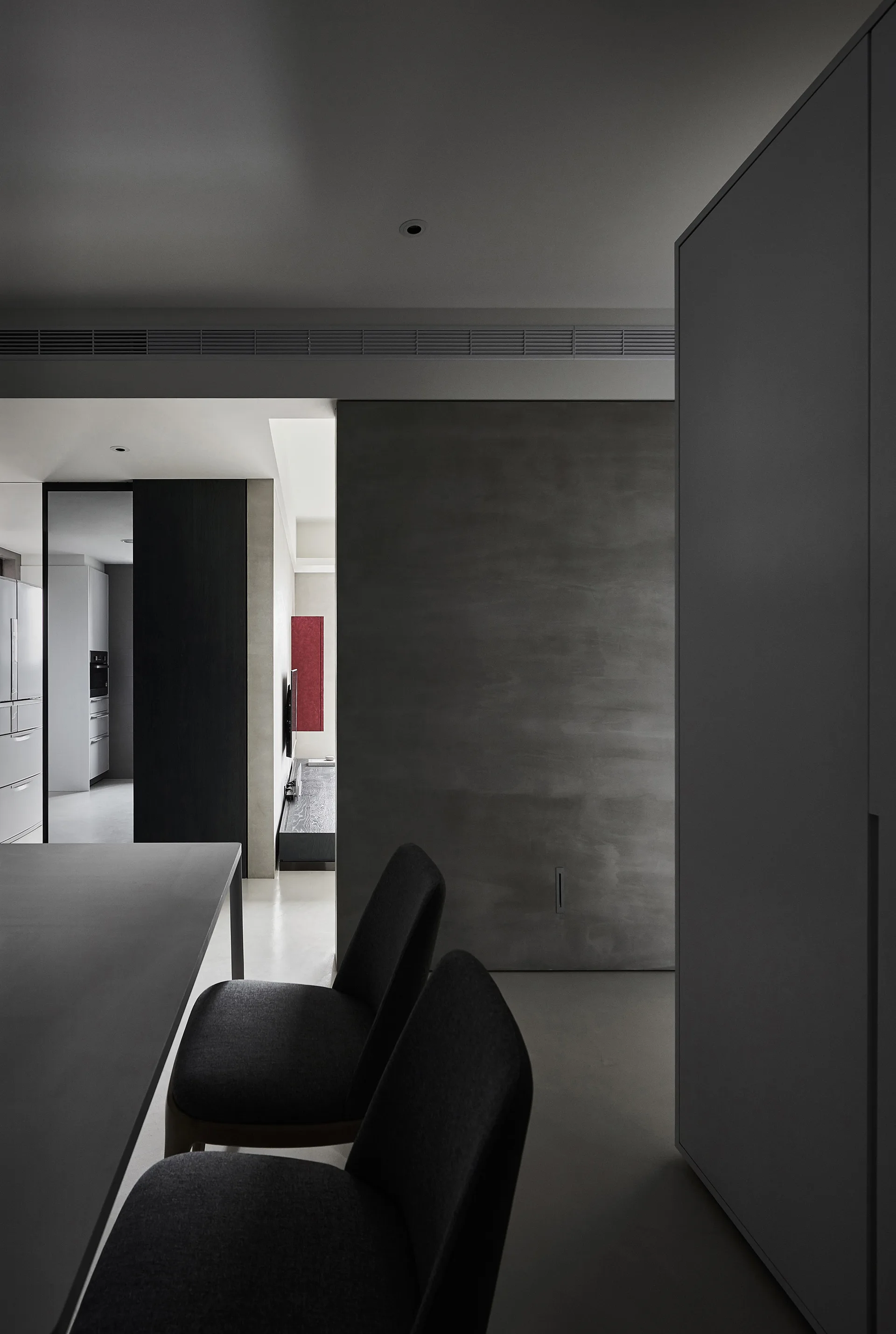 A minimalist dining area featuring dark upholstered chairs next to a grey concrete-textured wall, with a view into an adjacent kitchen.