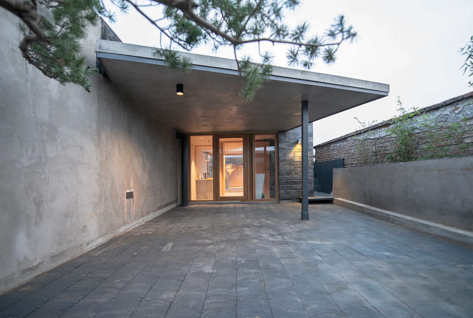 A modern minimalist concrete patio featuring dark textured floor pavers, a smooth raw concrete wall, and a warm wood-framed glass entryway.