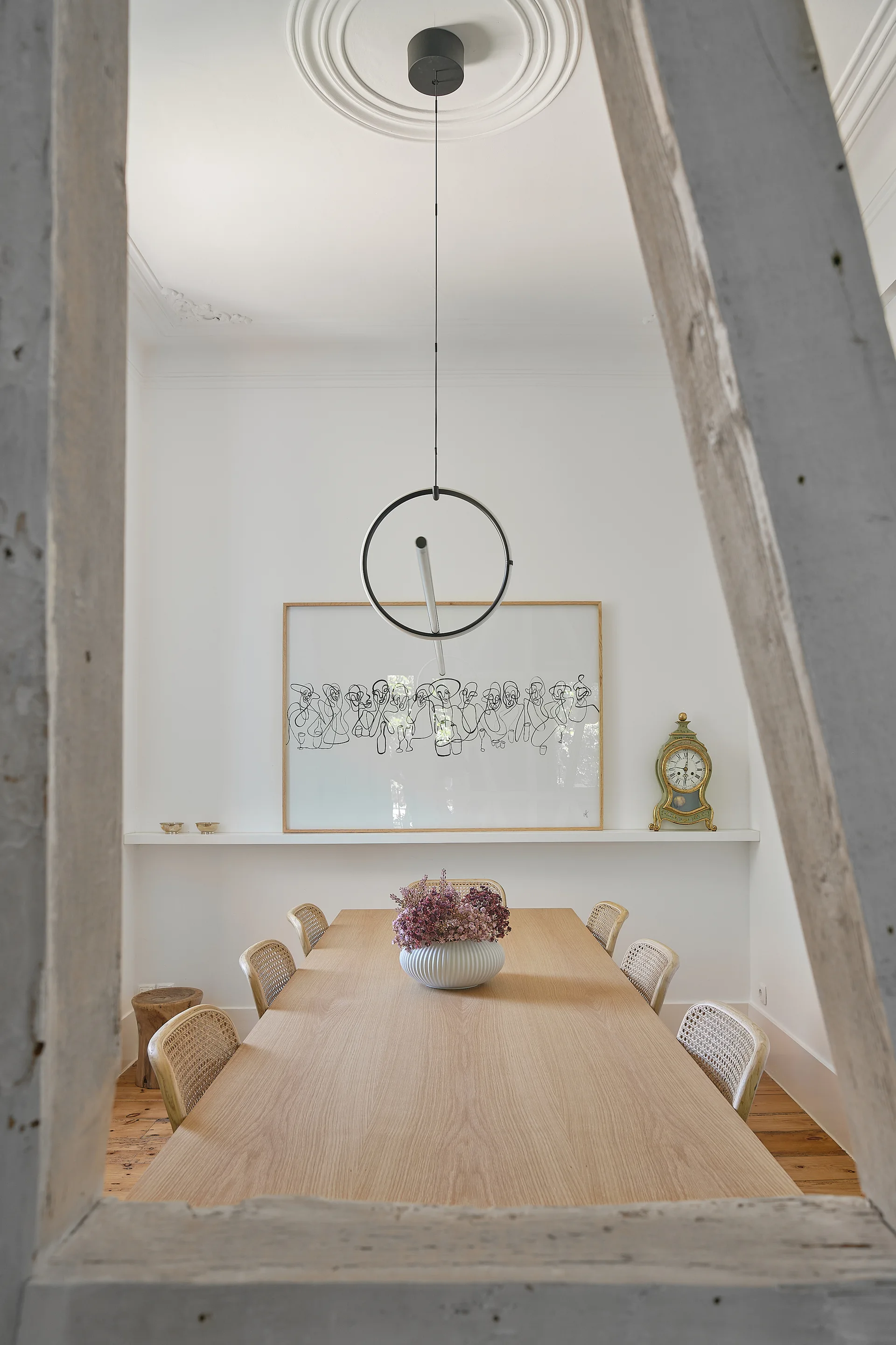 A minimalist dining room with a light wood table, rattan chairs, and a circular pendant light hanging from an ornate ceiling medallion.