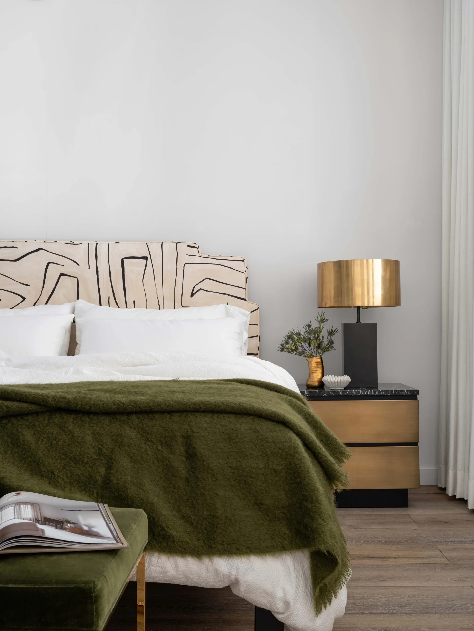 A bedroom featuring an upholstered headboard with an abstract black line pattern, a gold metal and black nightstand, and an olive green throw blanket.