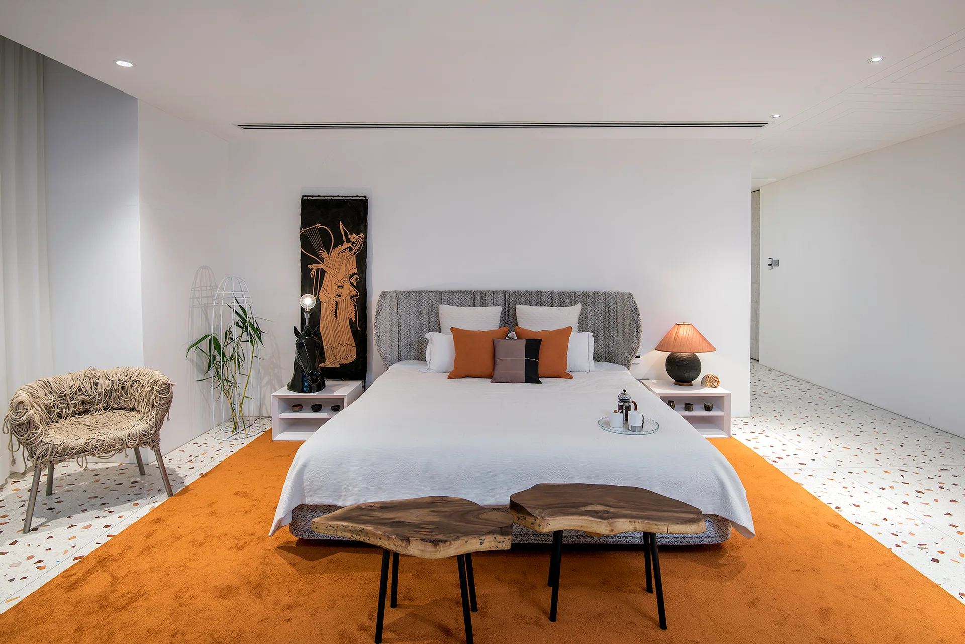 A contemporary bedroom features a bed with white bedding, a bright orange rug on a terrazzo floor, and two live-edge wood side tables at the foot.