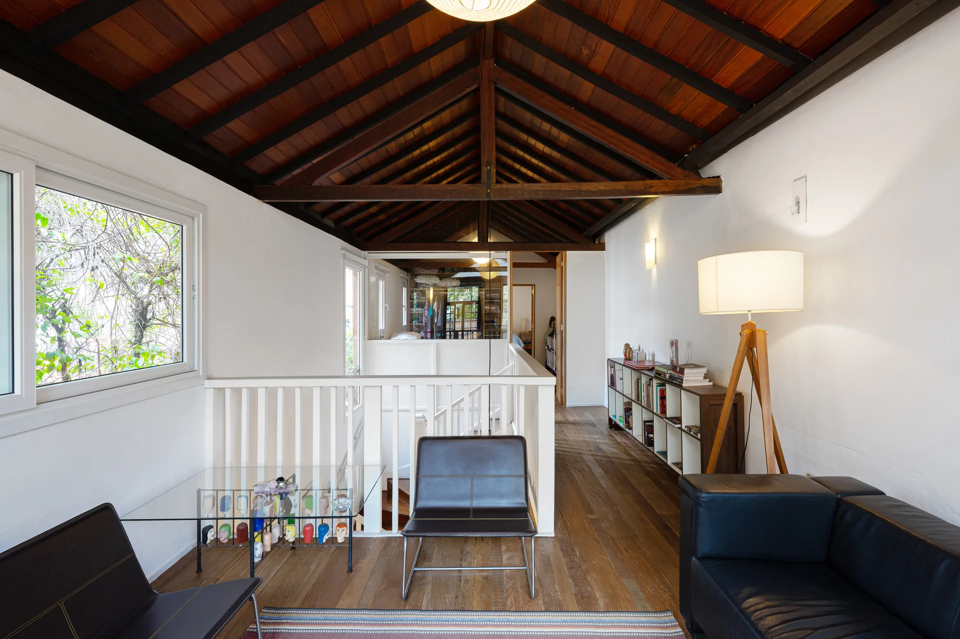 A home landing area features a vaulted timber ceiling, white walls, wooden floors, a white staircase railing, and black leather seating.
