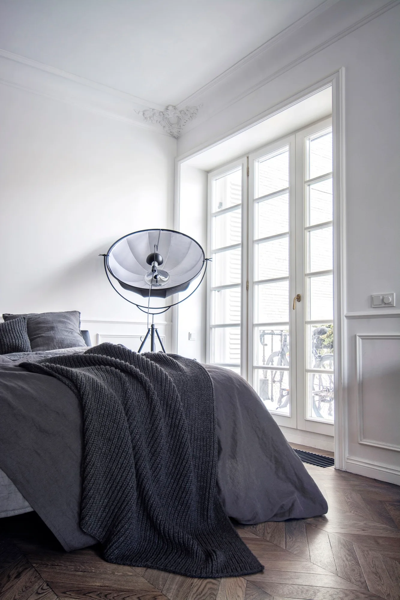 A minimalist bedroom features dark gray bedding, a large studio tripod floor lamp, and tall glass French doors opening to the outside.