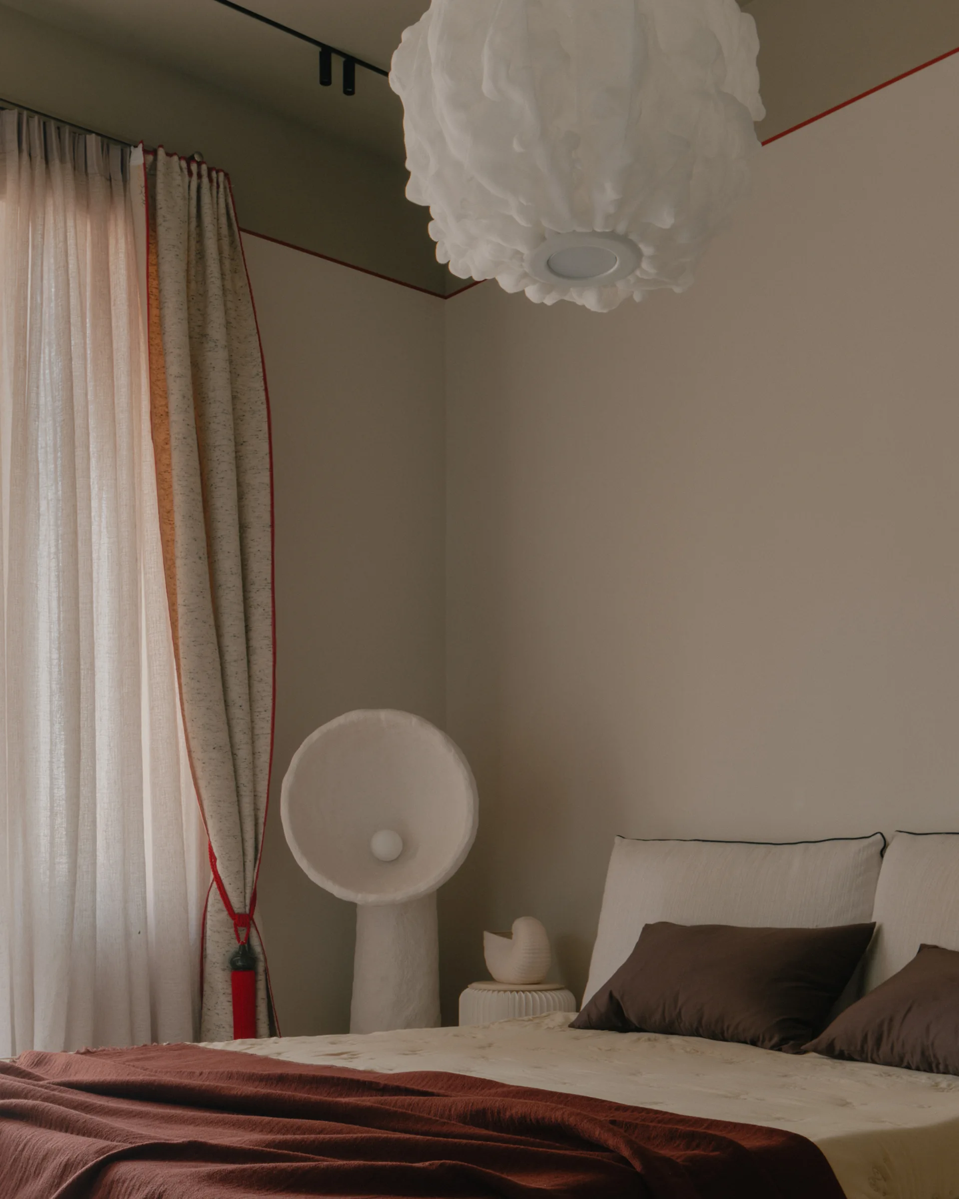 A minimalist bedroom features a bed with brown pillows, a sculptural white floor lamp, a cloud-like pendant light, and a thin red line on the wall.