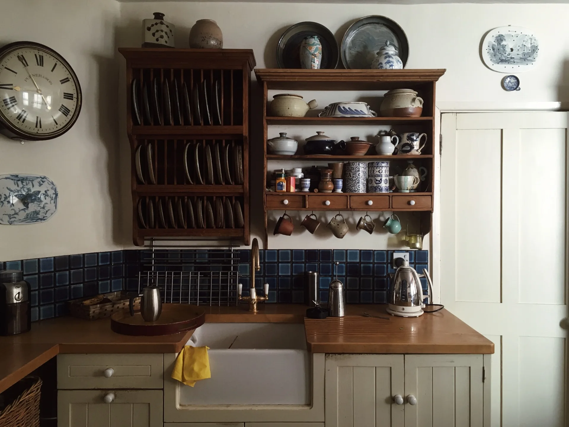 A rustic kitchen features a white farmhouse sink with a brass faucet, blue tile backsplash, and two wooden open plate racks on the wall.