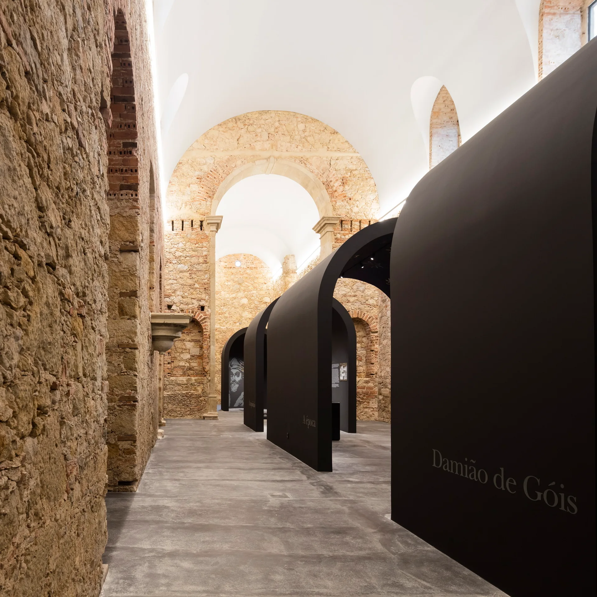 Smooth black curved exhibition partitions stand on a gray concrete floor inside a room with rough ancient stone walls and a vaulted ceiling.