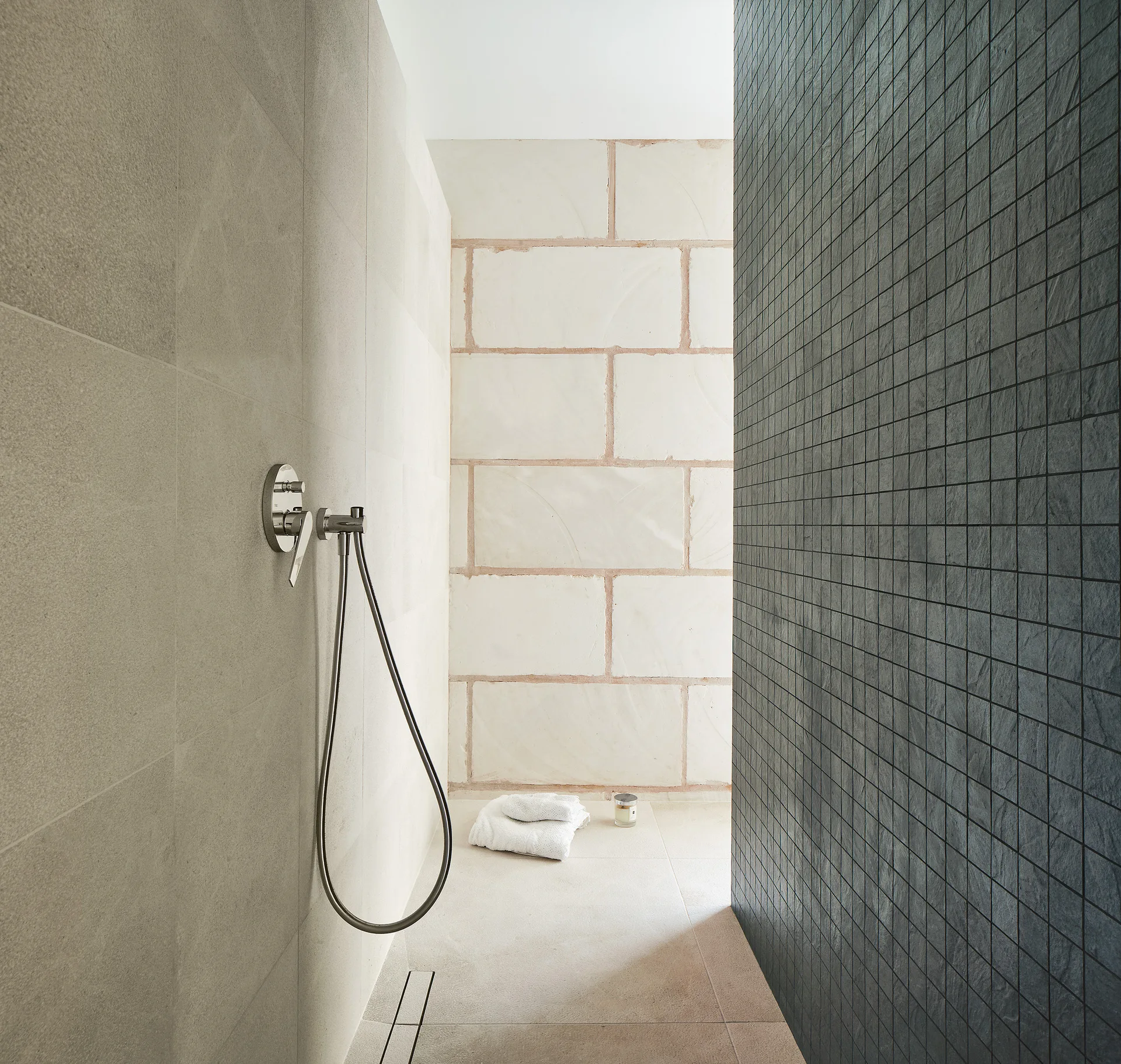 A minimalist walk-in shower featuring a dark charcoal mosaic tile wall contrasting with light beige large-format stone tiles and chrome fixtures.