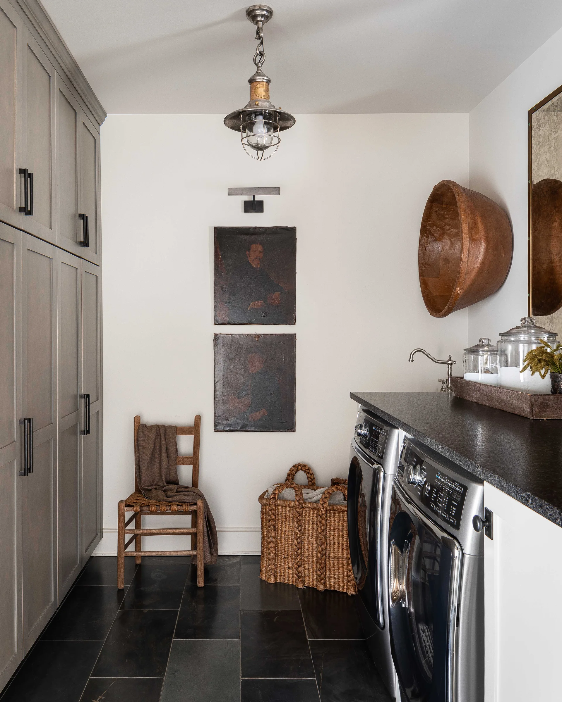 A laundry room featuring tall grey cabinets, dark slate flooring, a washer and dryer unit, and two stacked portrait paintings on the wall.