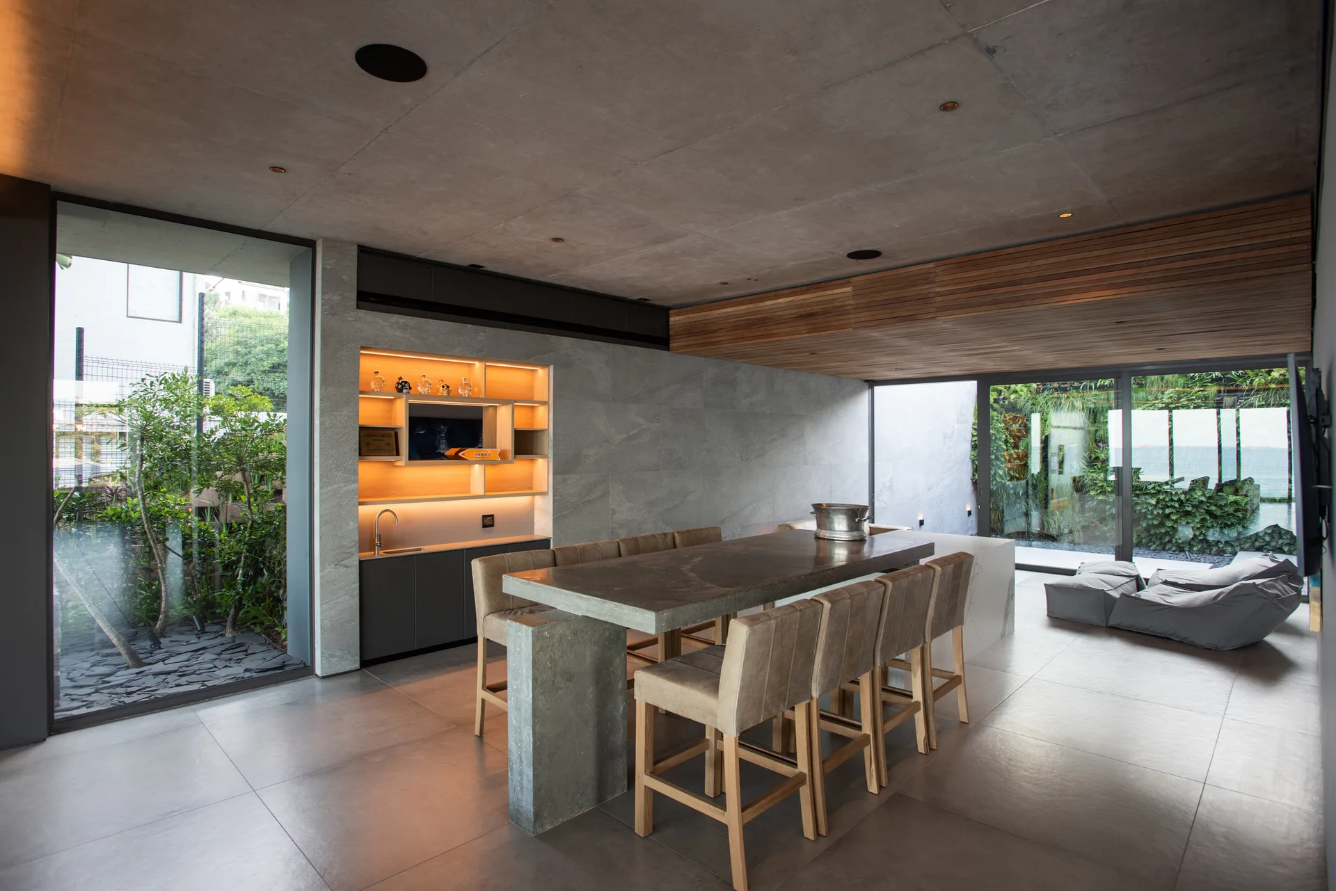 A modern dining area features a long concrete table surrounded by high-backed bar stools beneath a concrete ceiling and adjacent to sliding glass doors.