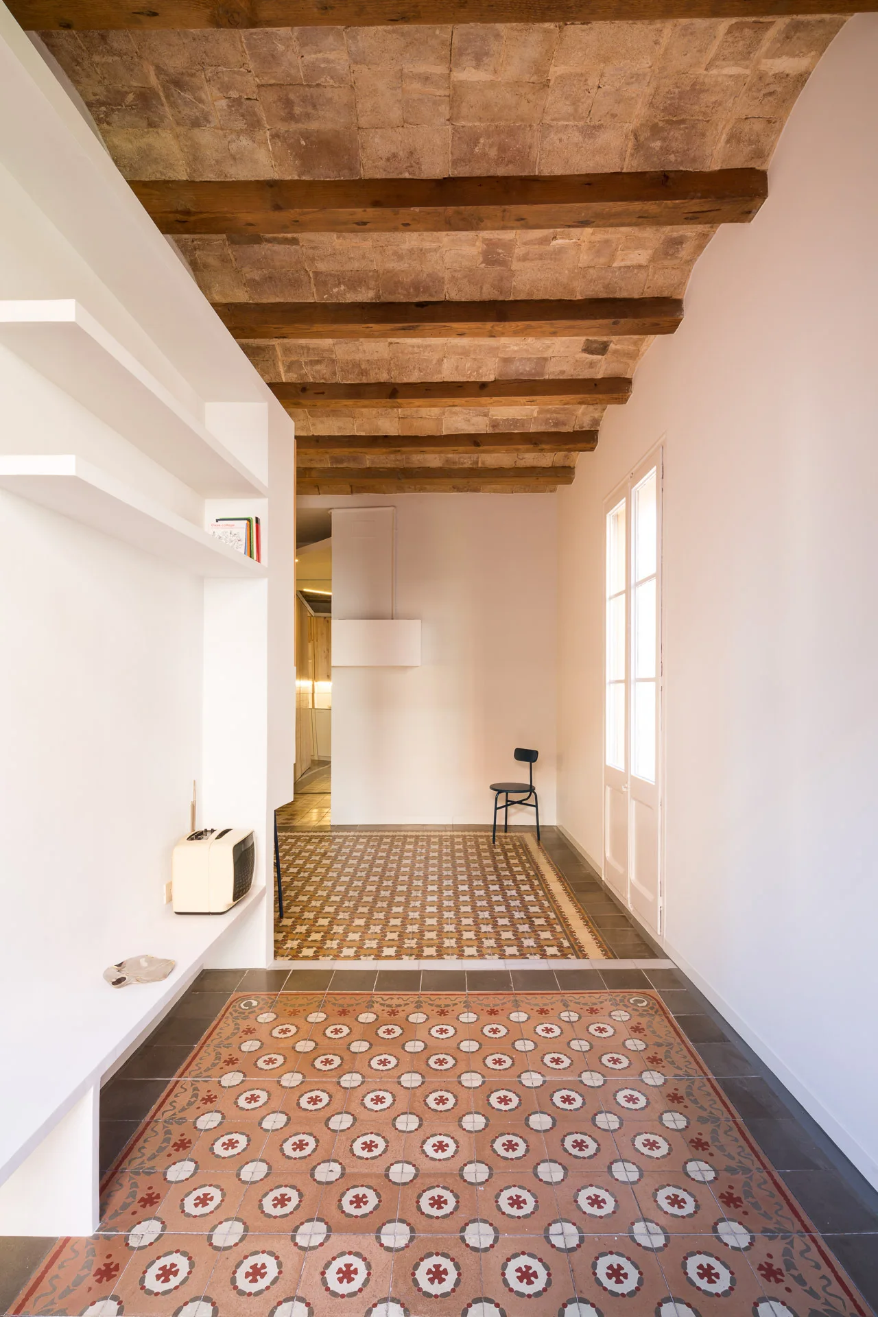 An airy hallway features a vaulted brick ceiling with wood beams, smooth white walls with built-in shelves, and a patterned terracotta tile floor.