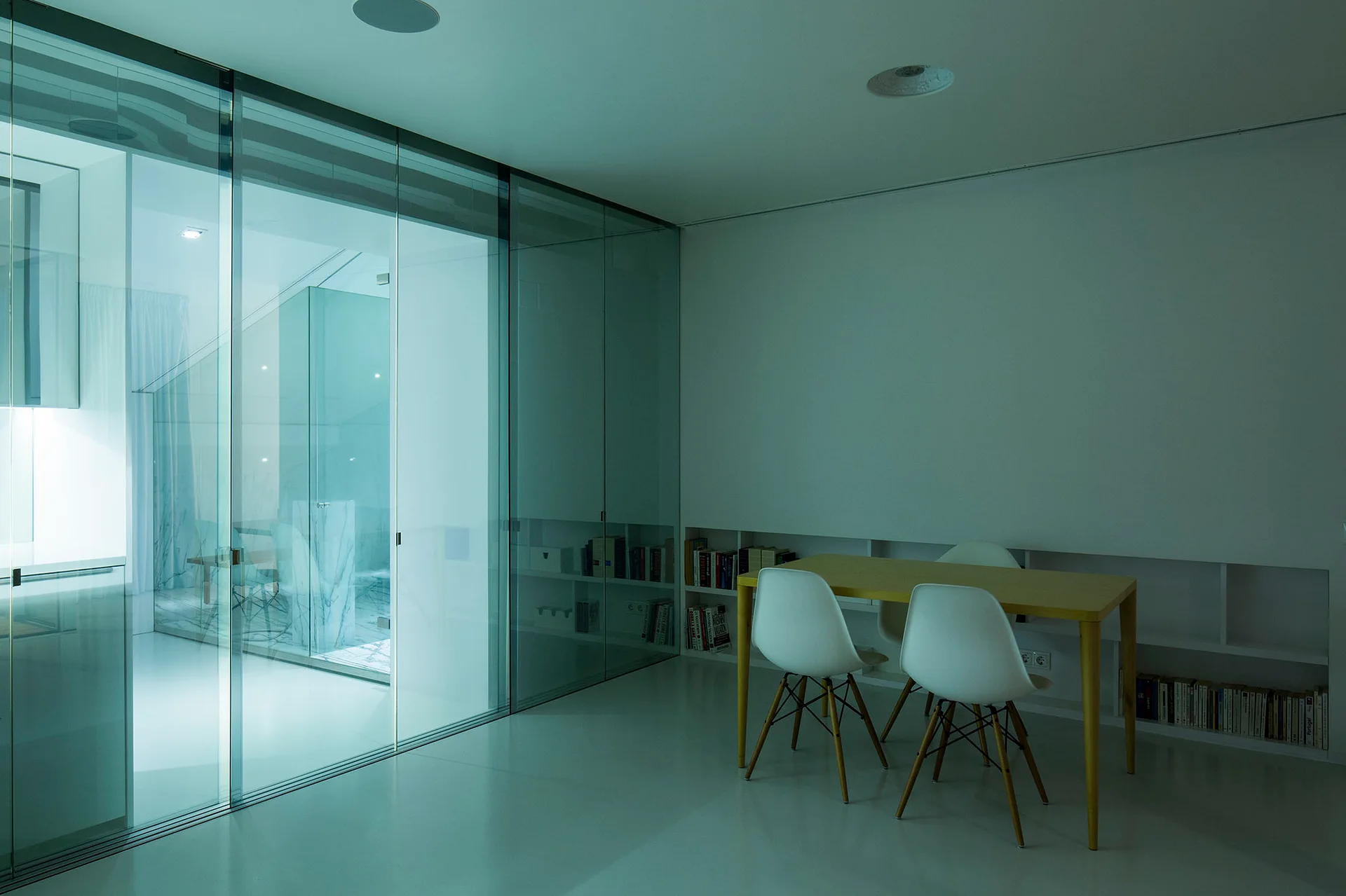A minimalist dining space features a light wood table, white molded chairs, a glass partition wall, and low recessed shelving filled with books.