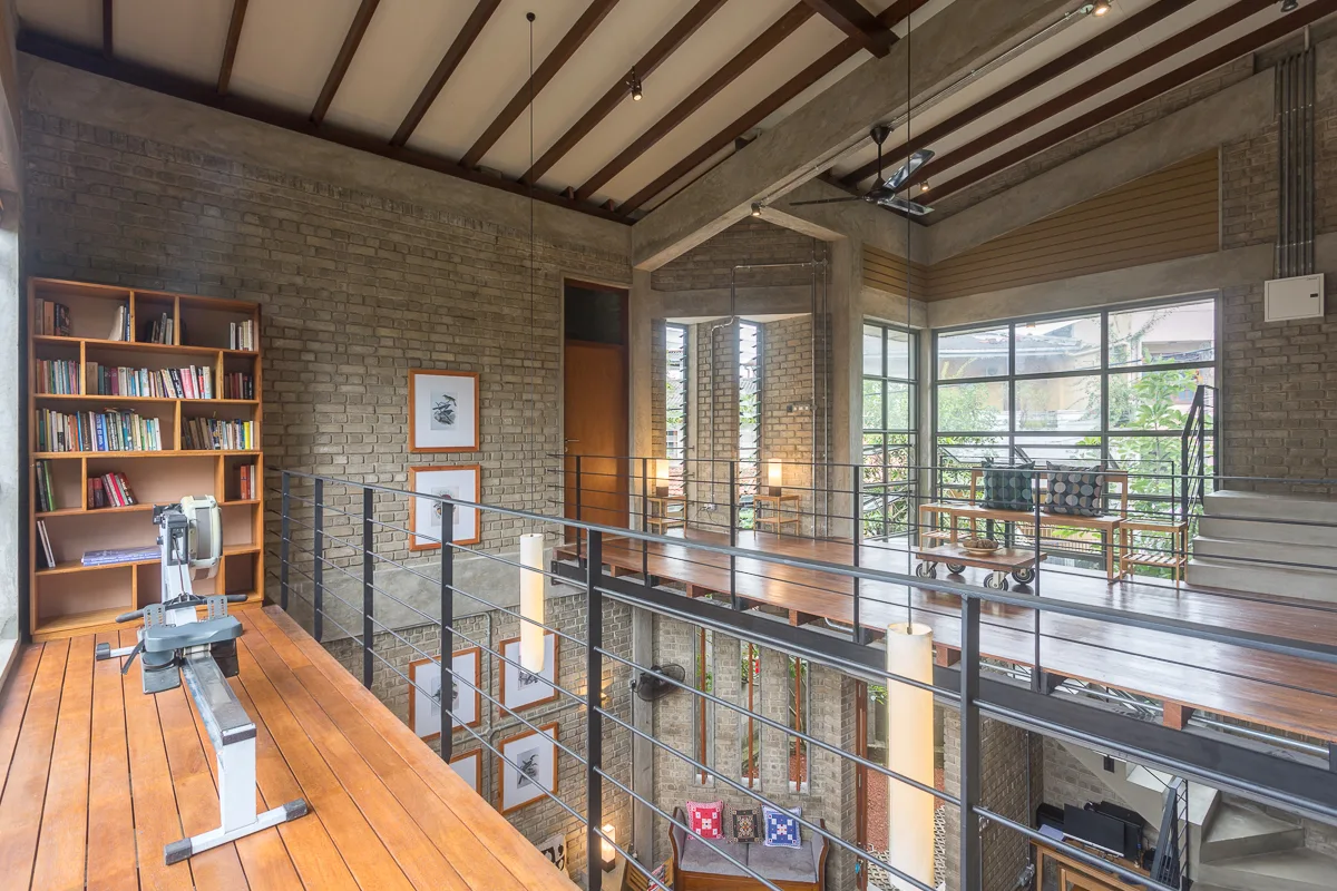 A mezzanine landing with warm wood floors features a tall bookshelf, a rowing machine, black steel railings, and textured grey-brown brick walls.