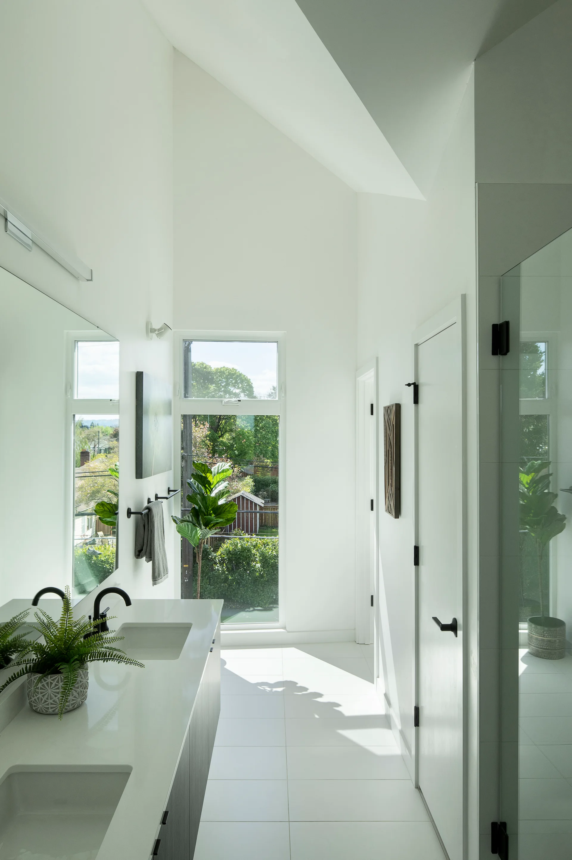 A bright bathroom features a double vanity with matte black faucets, white tile flooring, a large window, and a high vaulted ceiling.