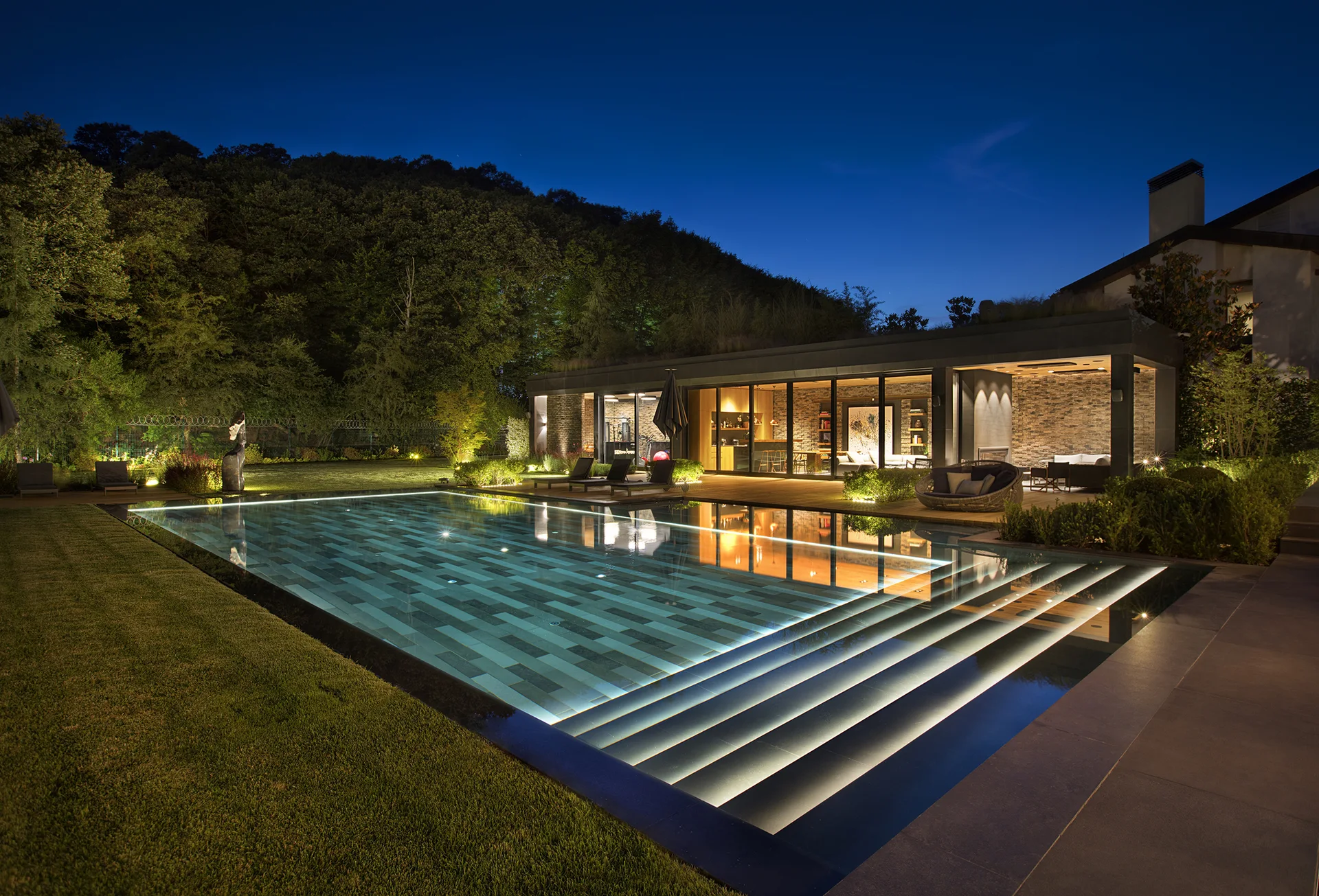 A modern rectangular swimming pool features illuminated stone steps alongside a wood deck and contemporary pool house at night.
