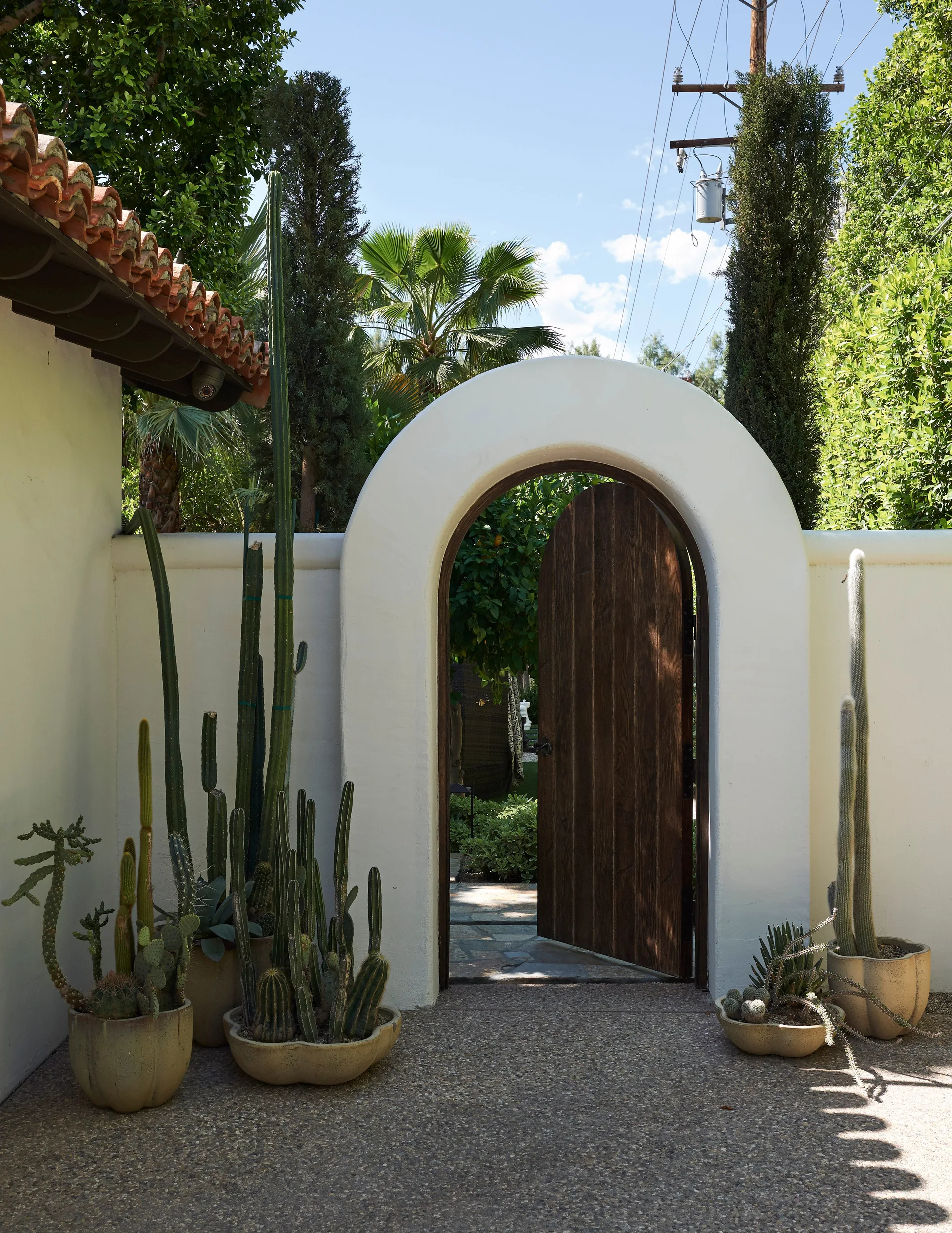 A white stucco wall features a curved archway with a dark wood door open to a lush garden, framed by potted cacti on a pebbled path.