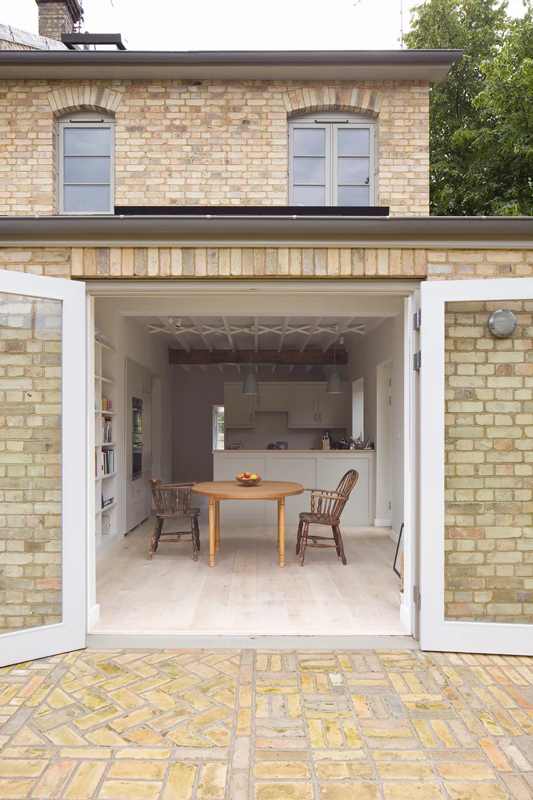 A view from a brick patio through open French doors into a kitchen with a round wooden table, two chairs, and exposed ceiling beams.