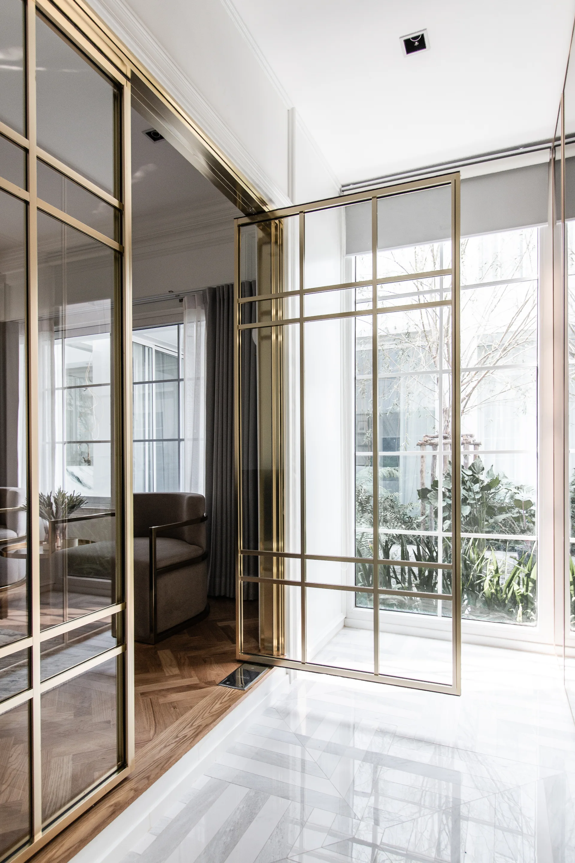 A large brass-framed glass pivot door stands partially open between a white tiled entryway and a room with herringbone wood flooring.