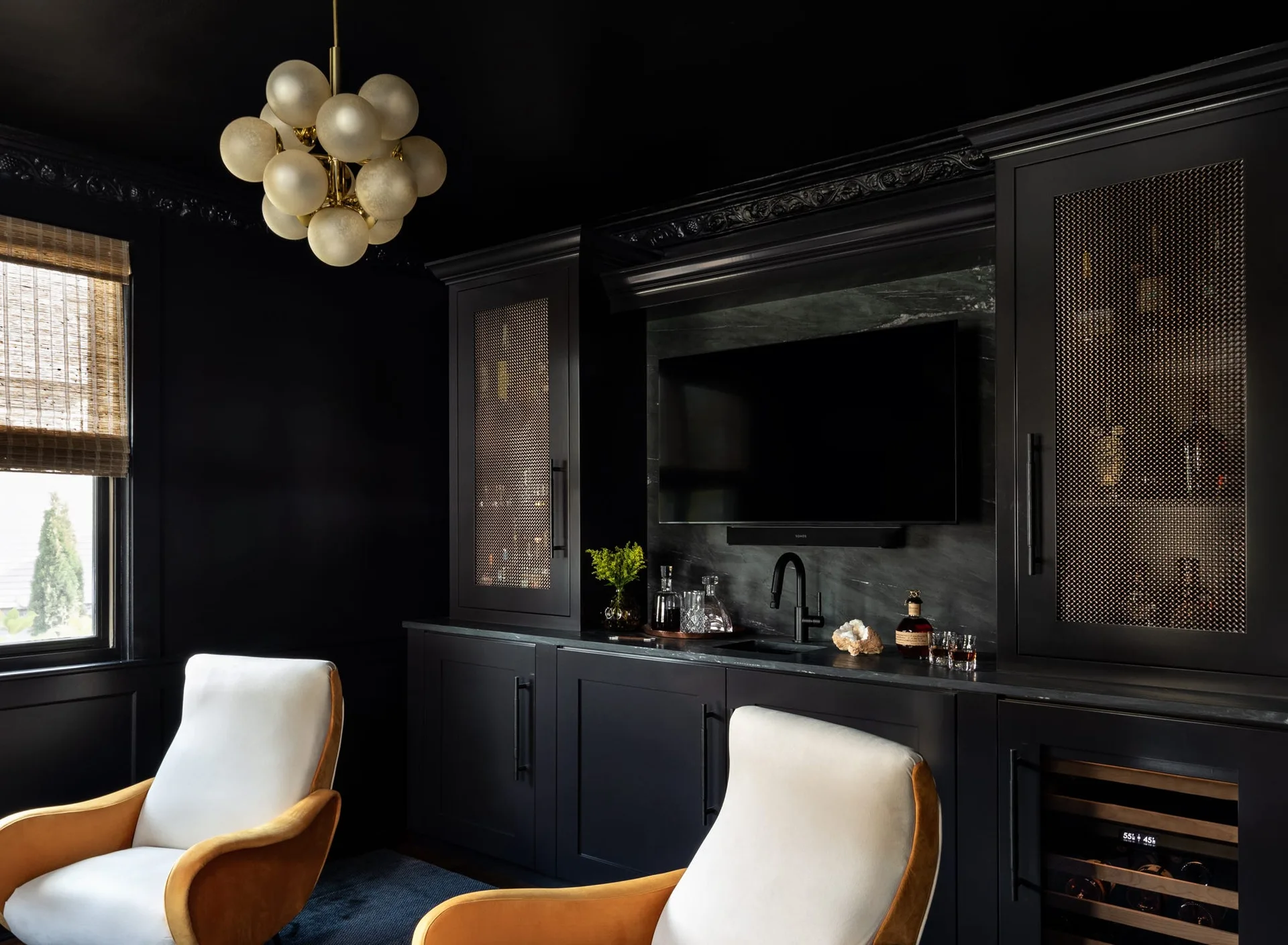 A moody home bar featuring matte black custom cabinetry with wire mesh door inserts, a black countertop, and a brass bubble chandelier overhead.