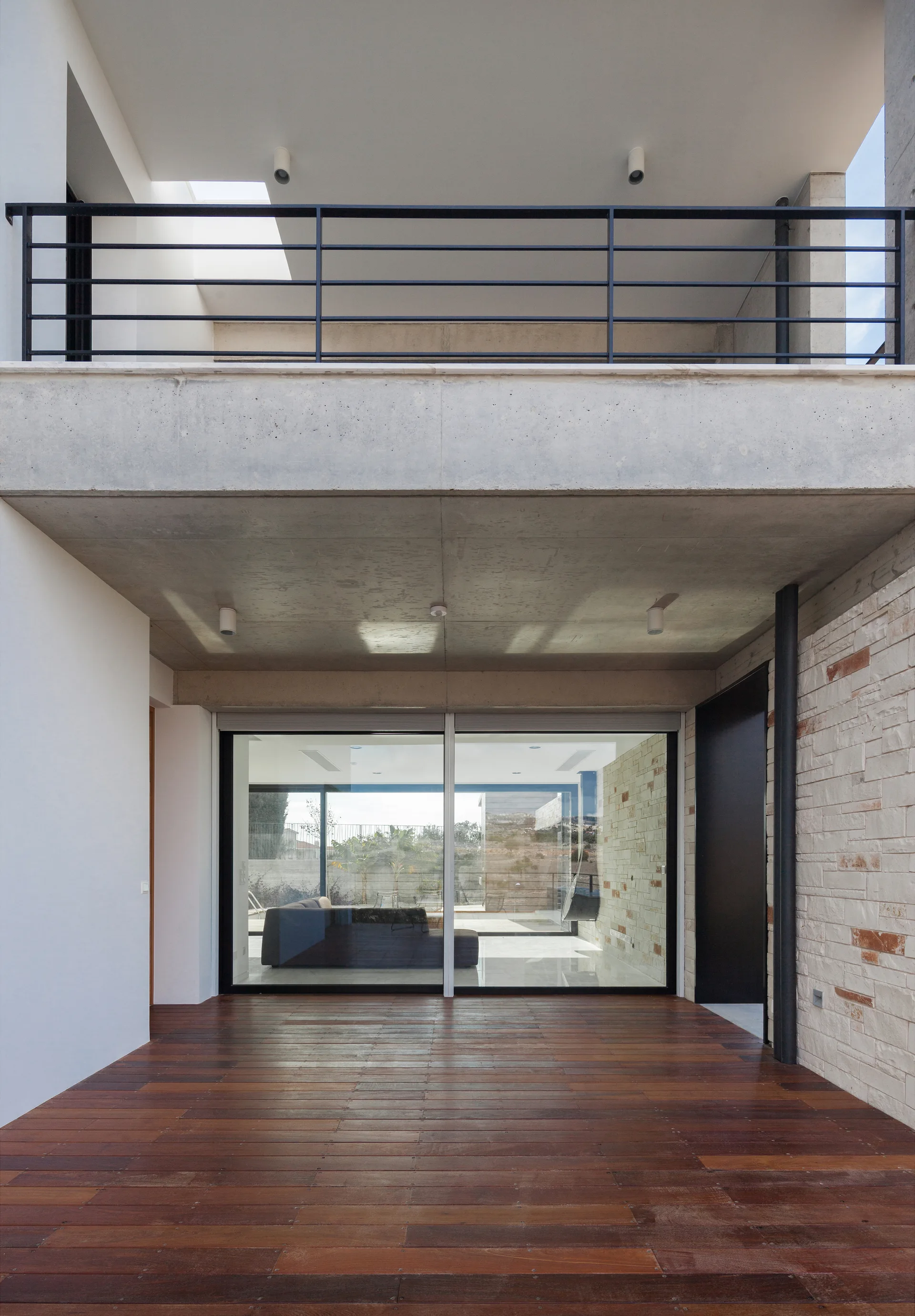 A covered modern patio features dark wood plank decking, an exposed concrete ceiling, and a large sliding glass door looking into a living room.