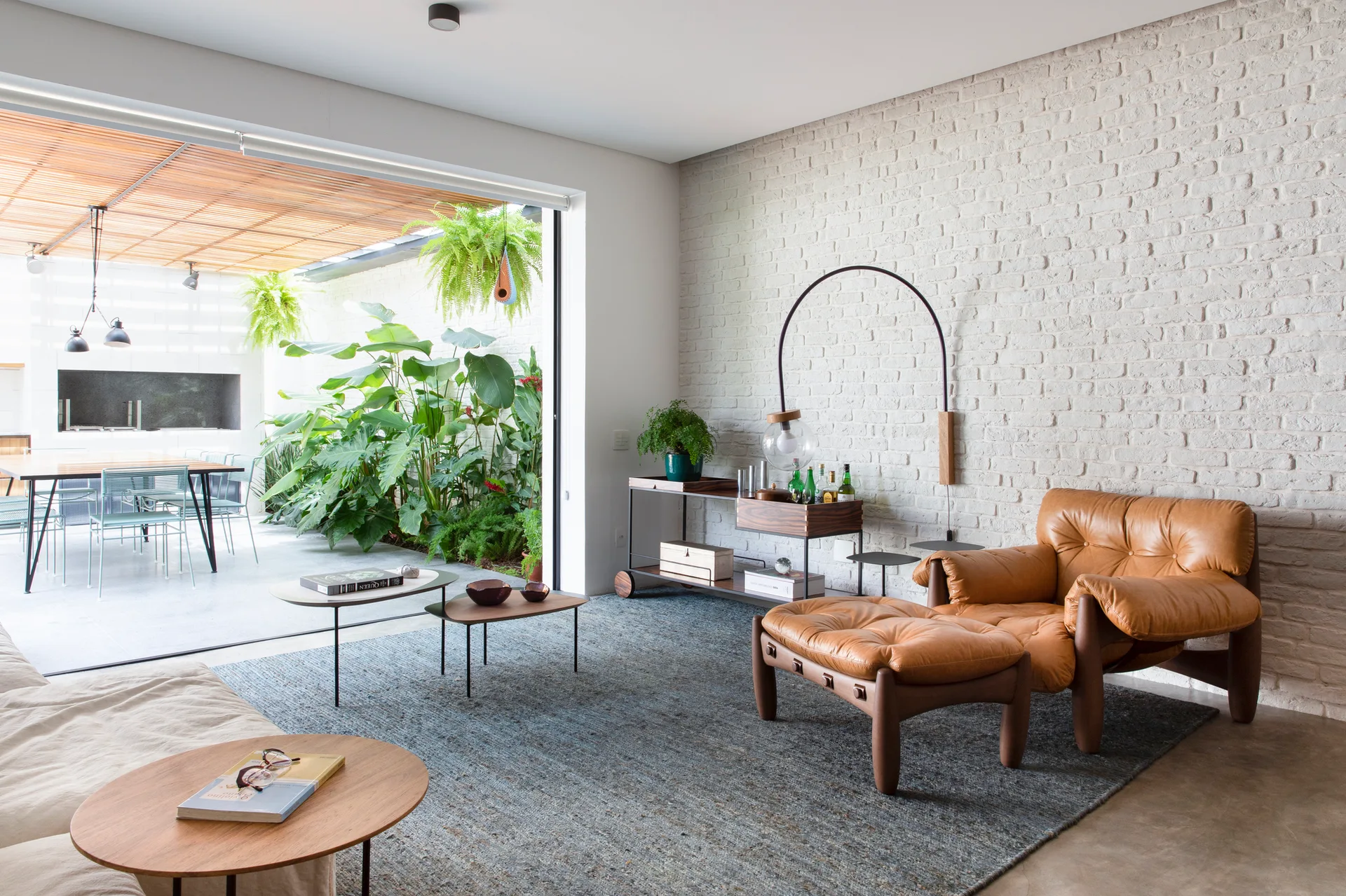 A modern living room features a tan leather lounge chair, grey area rug, and white brick wall, opening directly to a lush green outdoor patio.