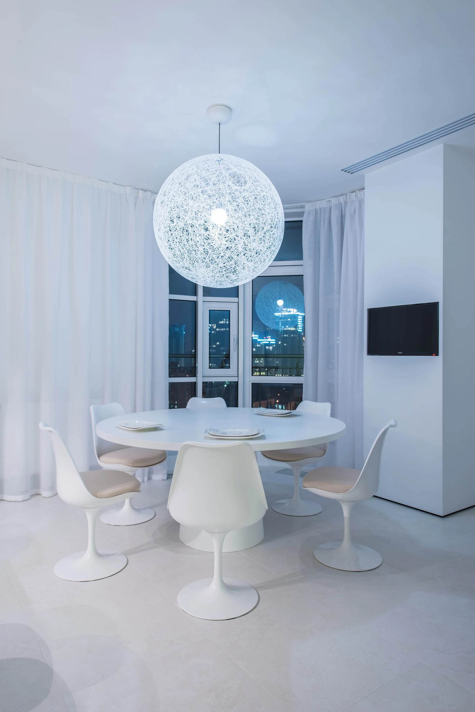 A minimalist white dining room features a round pedestal table, five tulip chairs, and a large spherical string pendant light hanging from the ceiling.