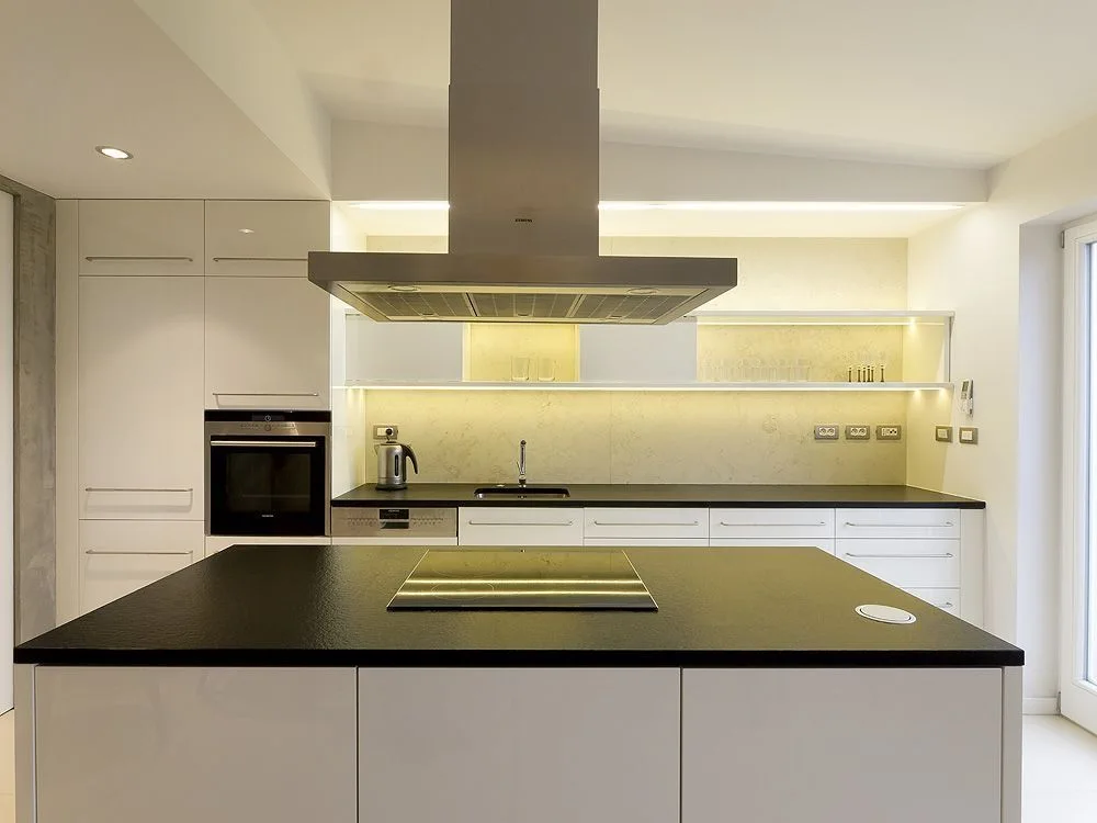A modern kitchen with a black-topped island, stainless steel range hood, white flat-front cabinetry, and a backlit open shelf.